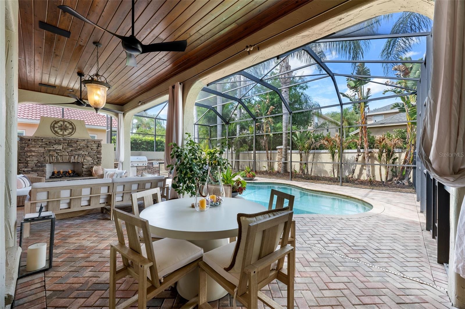 Covered lanai — Wood plank ceiling, ceiling fans, outdoor dining area, stone fireplace, and pool view.