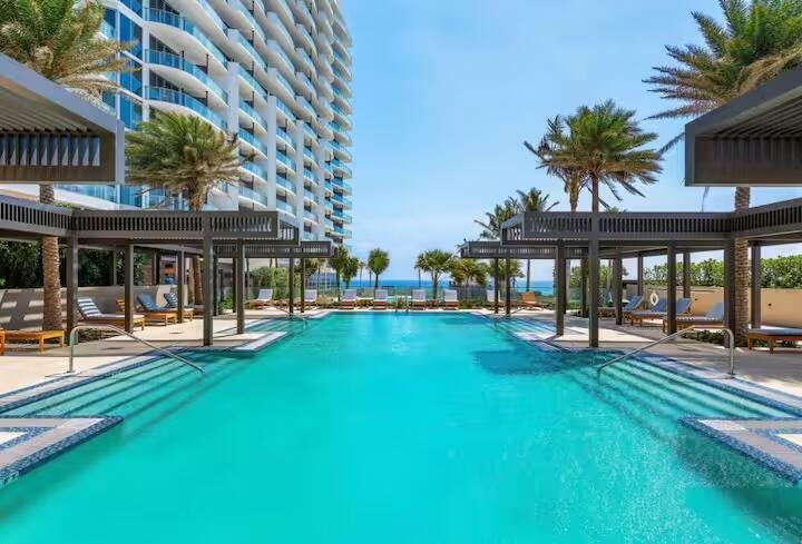 Expansive swimming pool with integrated steps, surrounded by lounging areas under pergolas, palm trees, and a multi-story building