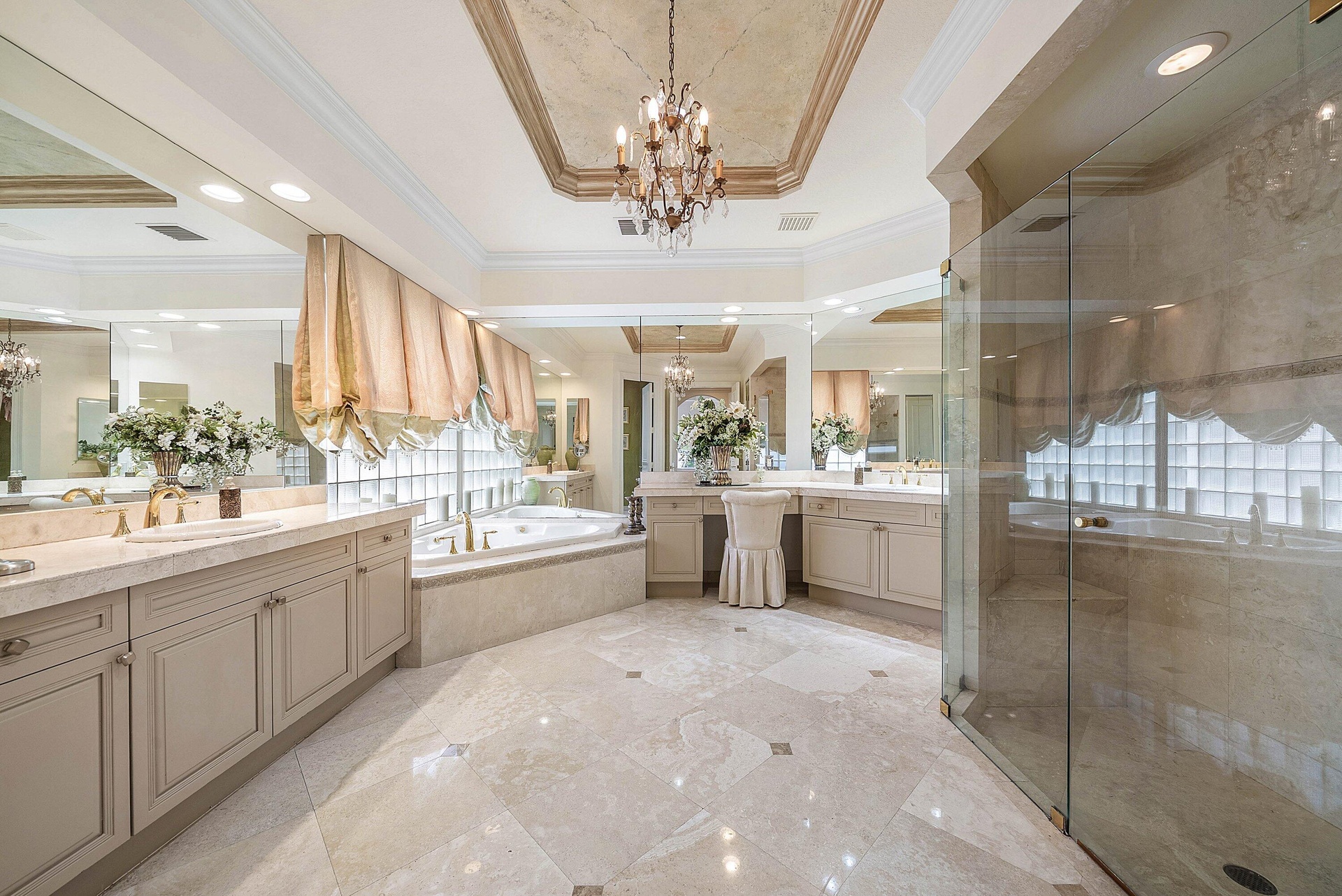 Marble flooring with accent details, dual vanities with integrated sinks, recessed jetted bathtub, glass-enclosed shower, and a decorative tray ceilin
