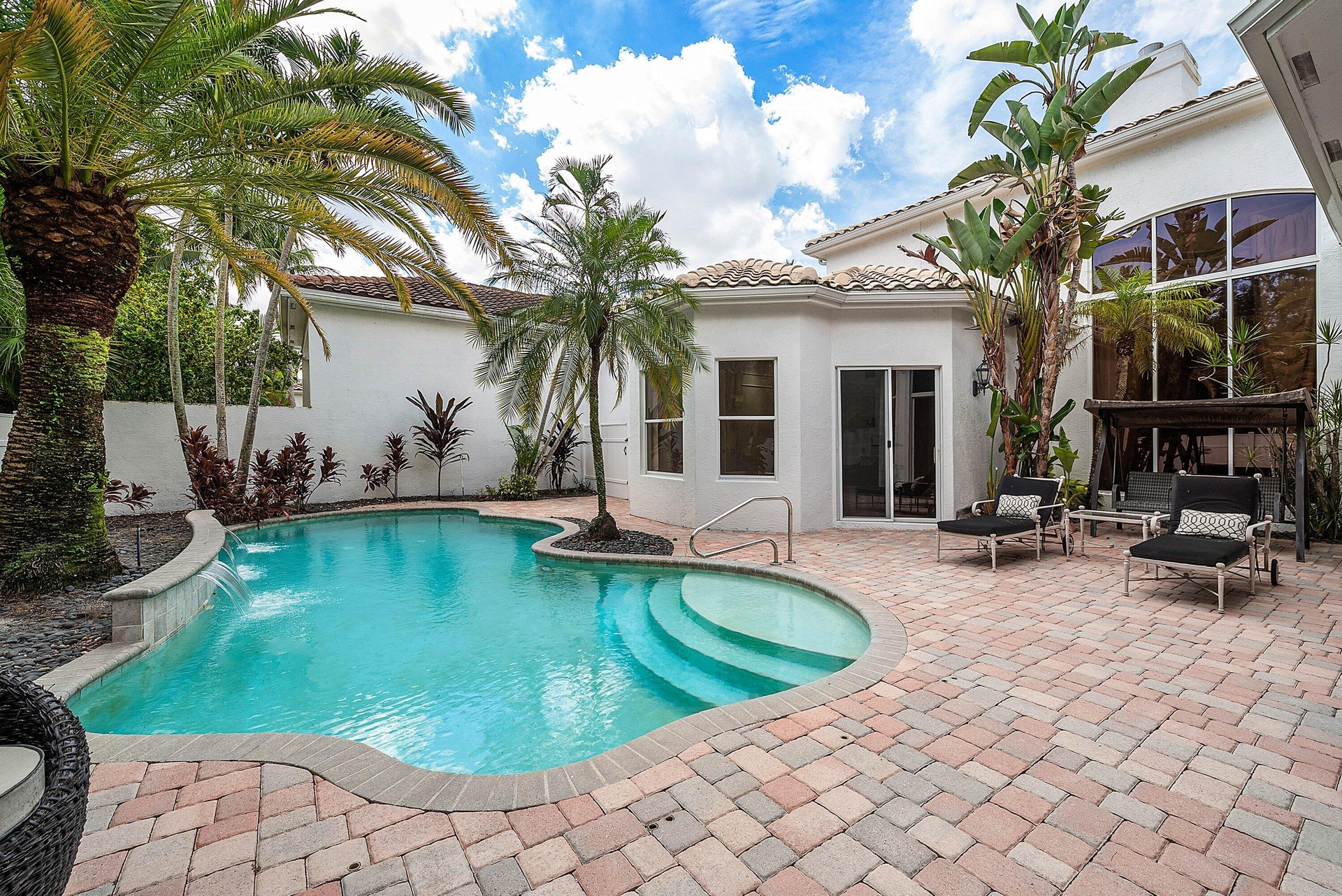 Curved swimming pool with waterfall feature, surrounded by a multi-tone paver patio and tropical landscaping