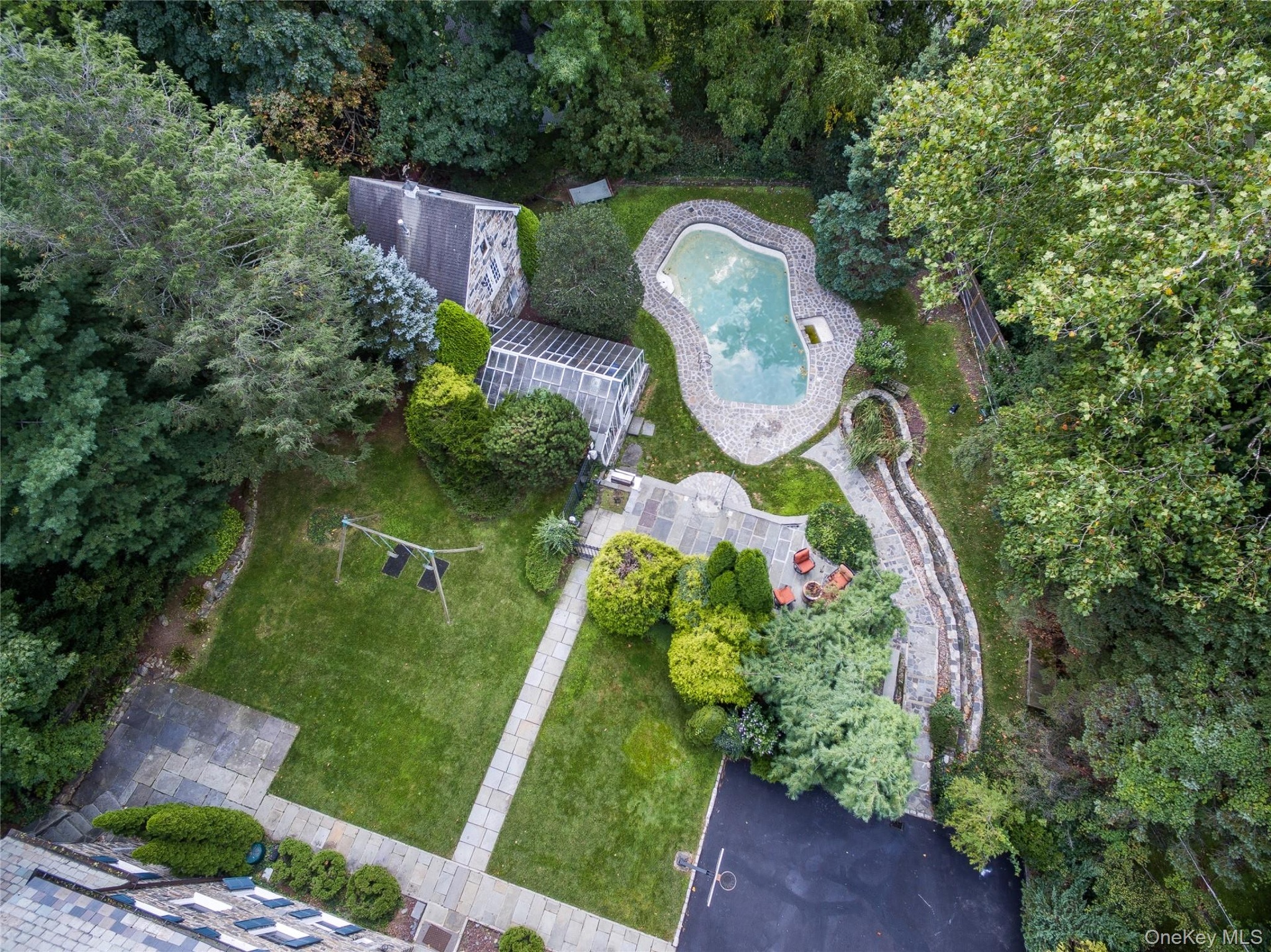 Aerial view of an expansive residential property featuring a custom-shaped swimming pool with integrated spa, manicured lawn, mature tree-lined perime