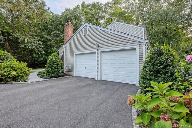 Side entry two-car garage with panel doors, asphalt driveway, brick chimney, and mature perimeter landscaping