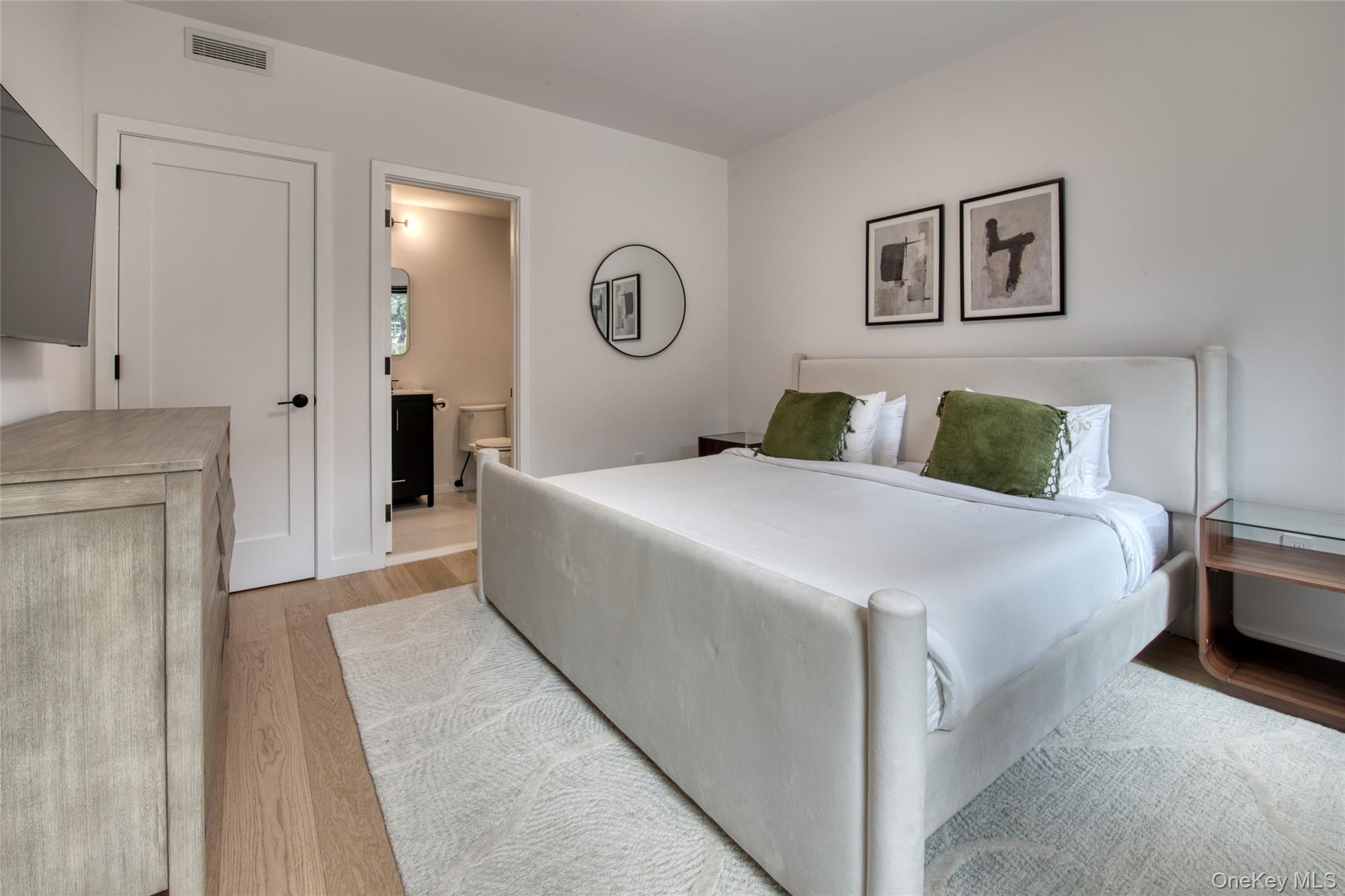 Light wood-finish flooring, white walls, and a private bath featuring a dark wood vanity and round mirror