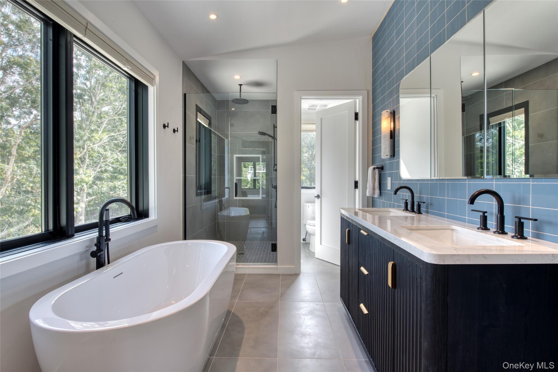 Freestanding soaking tub with black fixture, glass-enclosed shower, dual vanity with undermount sinks, and blue tiled accent wall