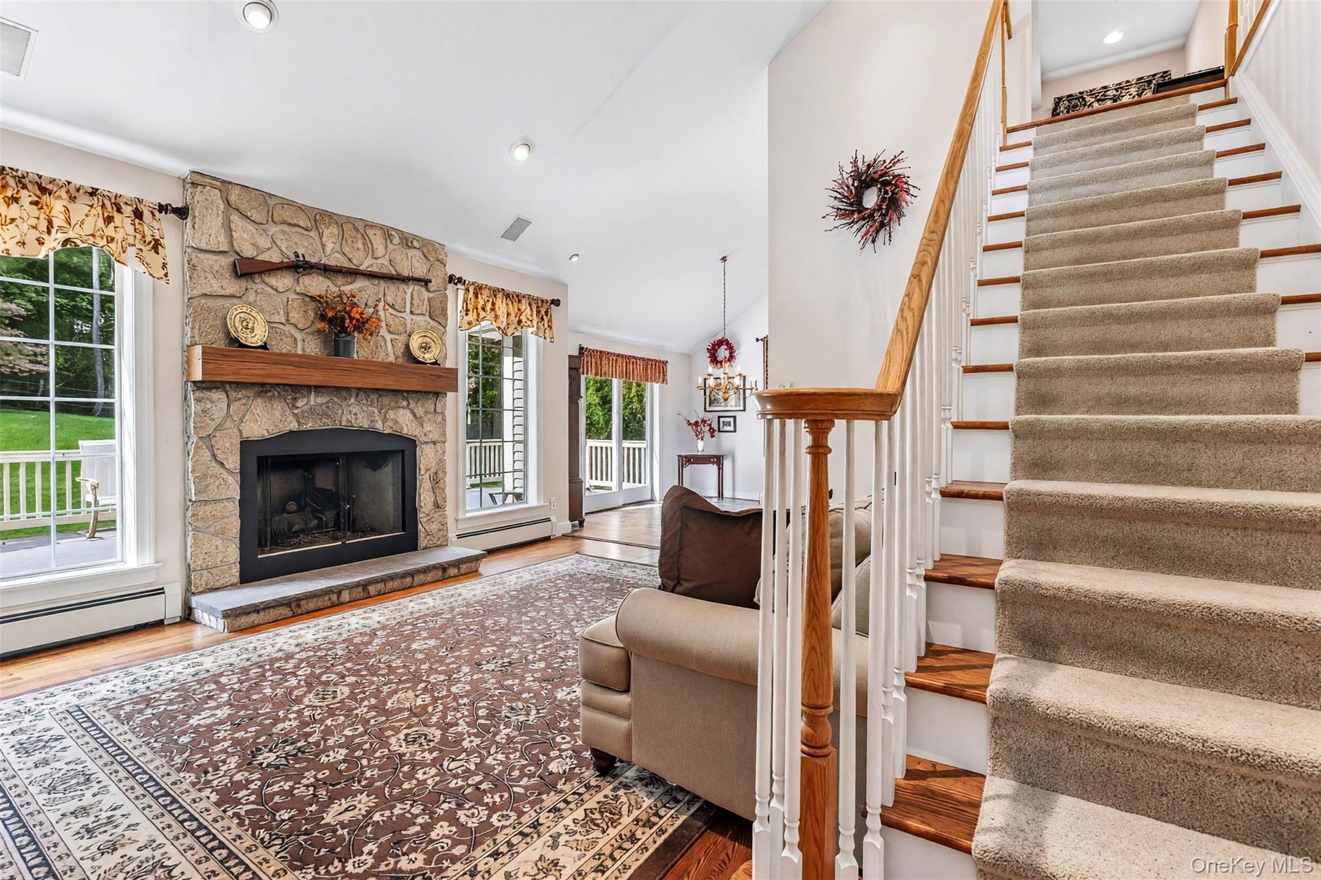 Stone-clad fireplace with a wood mantle, hardwood flooring, and a carpeted staircase with a wood handrail