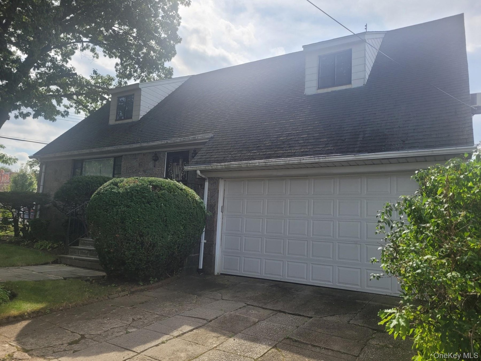 Stone-clad exterior with dormer windows, attached garage, concrete driveway, and established landscaping