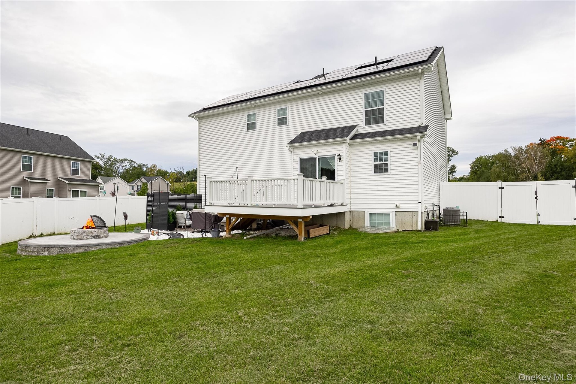 White vinyl siding exterior with solar panels, a private fenced yard, a large wood deck, and a circular stone fire pit