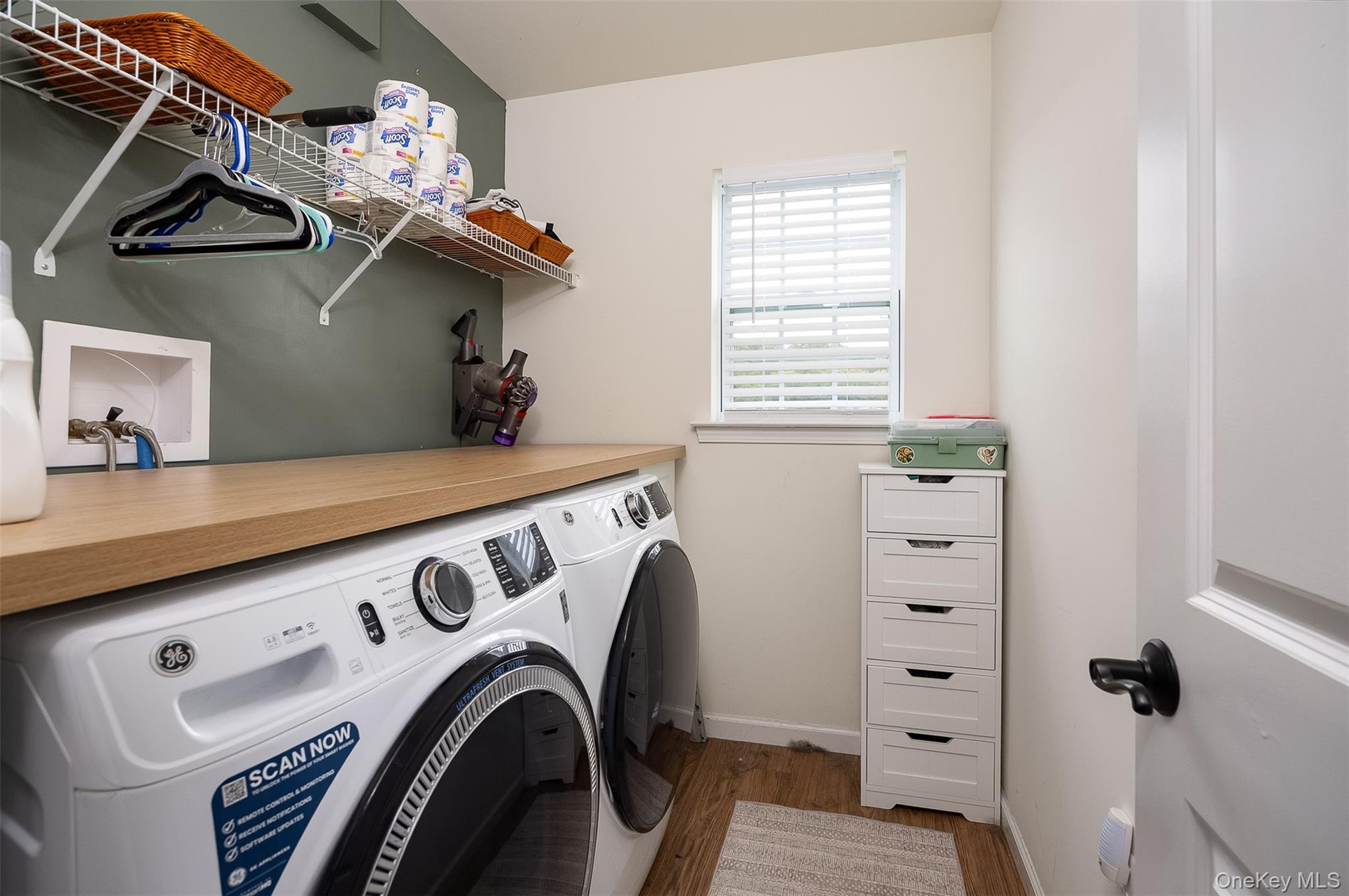 Laundry room with light wood-finish countertop, window with blinds, wire shelving, and a white paneled door with black hardware