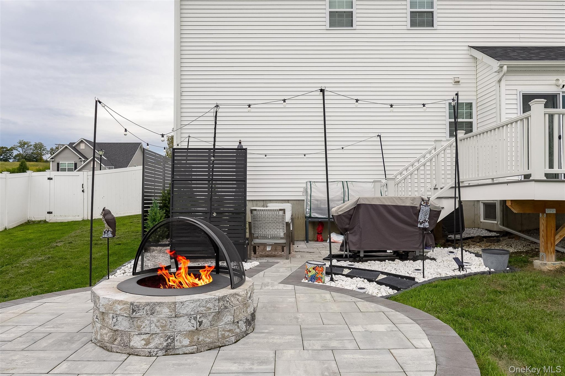 Stone-clad fire pit, paver patio, string lighting, white vinyl fencing, and white siding