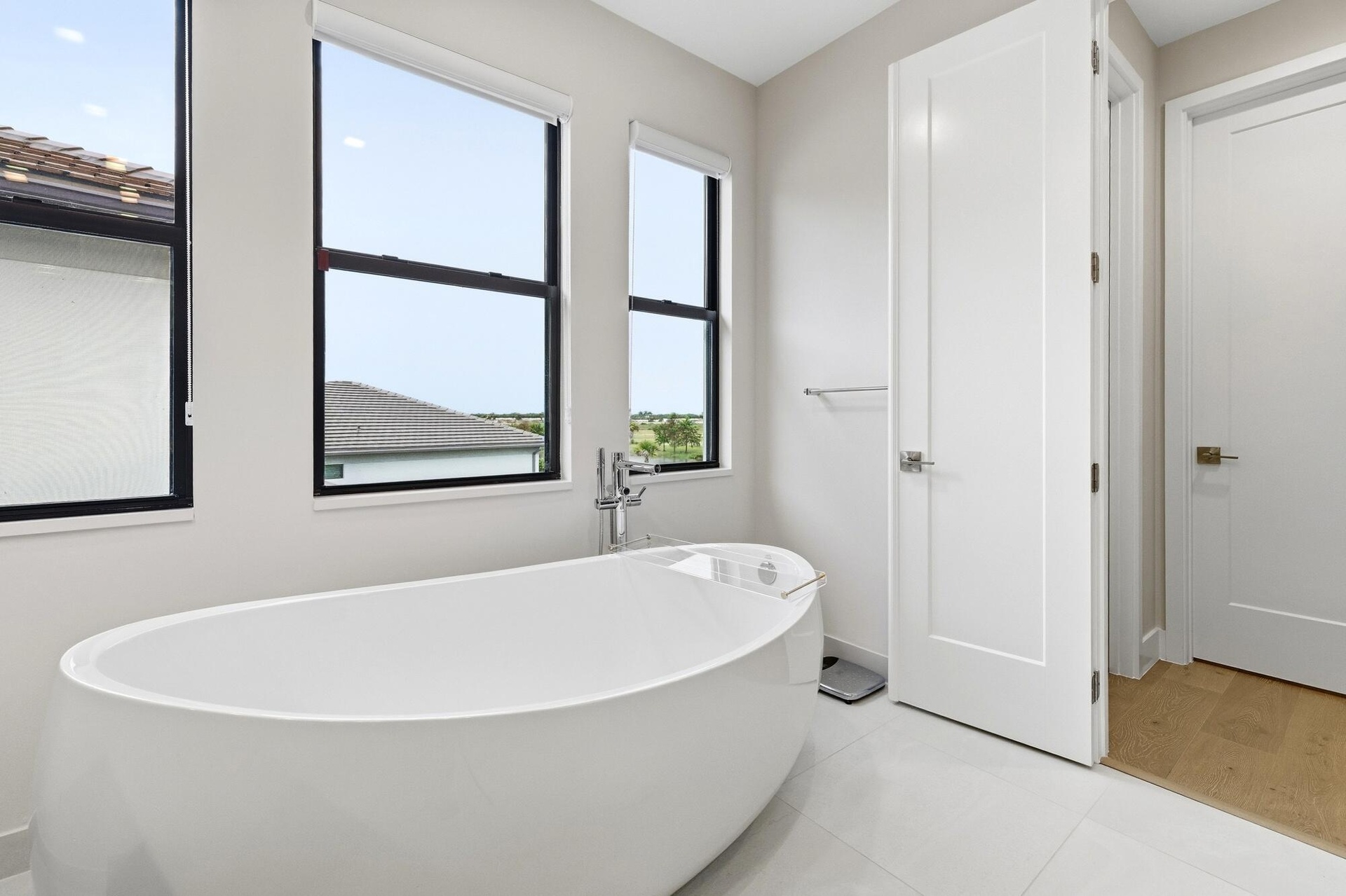 Freestanding soaking tub with floor-mounted faucet, complemented by large windows, white paneled doors, and light tile flooring
