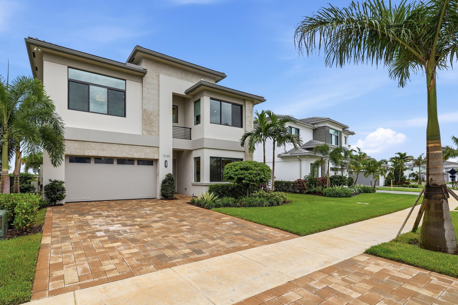 Modern architectural exterior with a stucco and stone facade, two-car garage, paver driveway, and mature landscaping