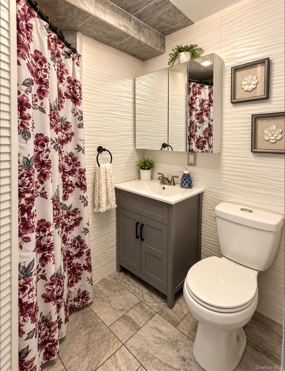 Wavy-textured wall tile, gray vanity with double cabinet doors, integrated sink, mirrored medicine cabinet, and large-format tile flooring