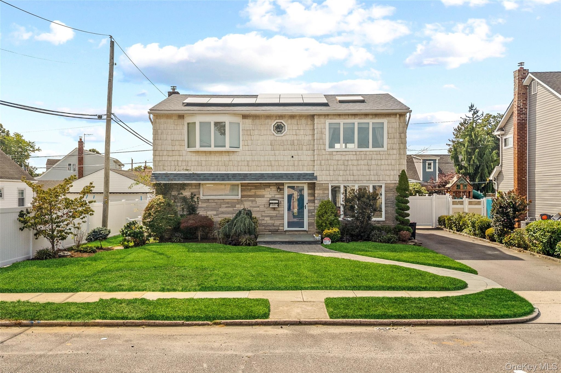 Two-story residence featuring a shingled exterior and stone facade, solar panels, a paved driveway, and a front lawn with a concrete pathway