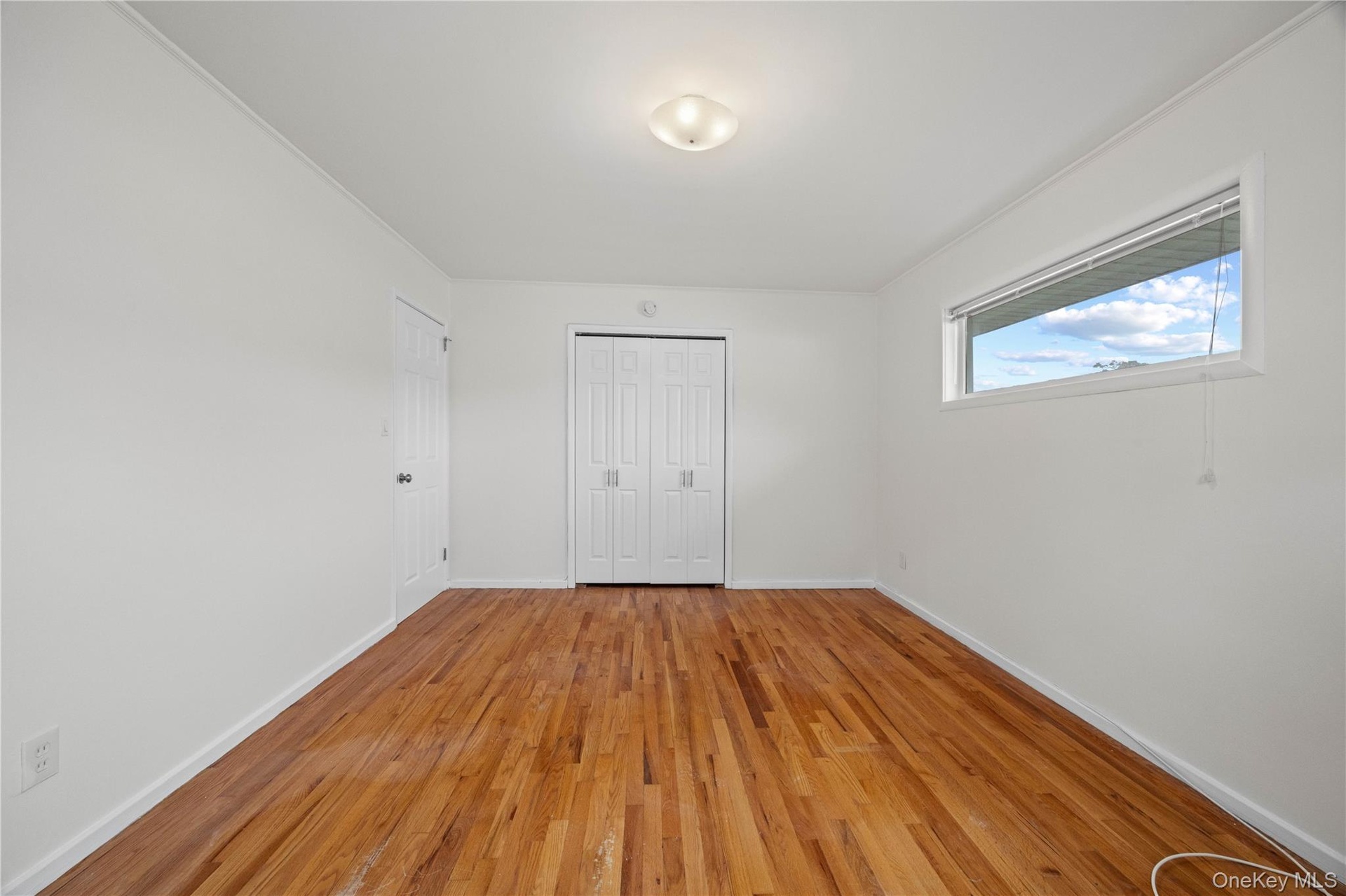 Hardwood flooring, white walls, flush mount ceiling light, horizontal window with roller shade, bi-fold closet doors