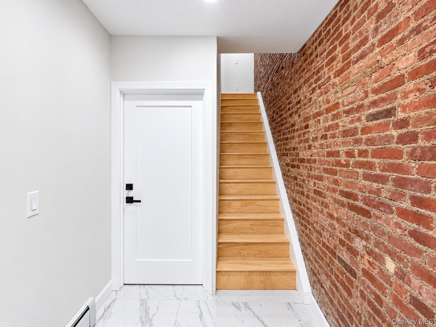Exposed brick accent wall, wood staircase with white risers, white panel door with matte black hardware, recessed ceiling lighting, and white tile flo