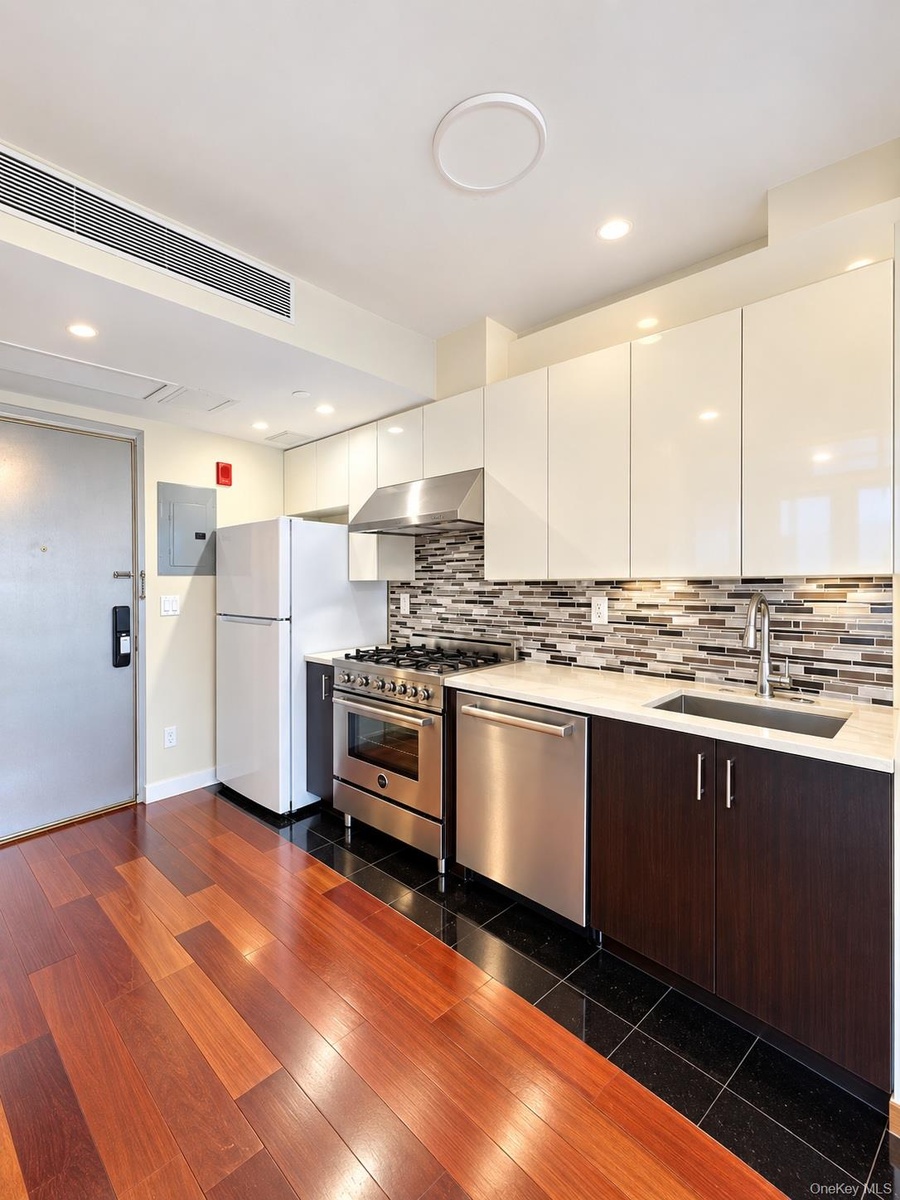 Gleaming wood-finish flooring, stainless steel appliances, white flat-panel cabinetry, a mosaic tile backsplash, and a light-toned countertop