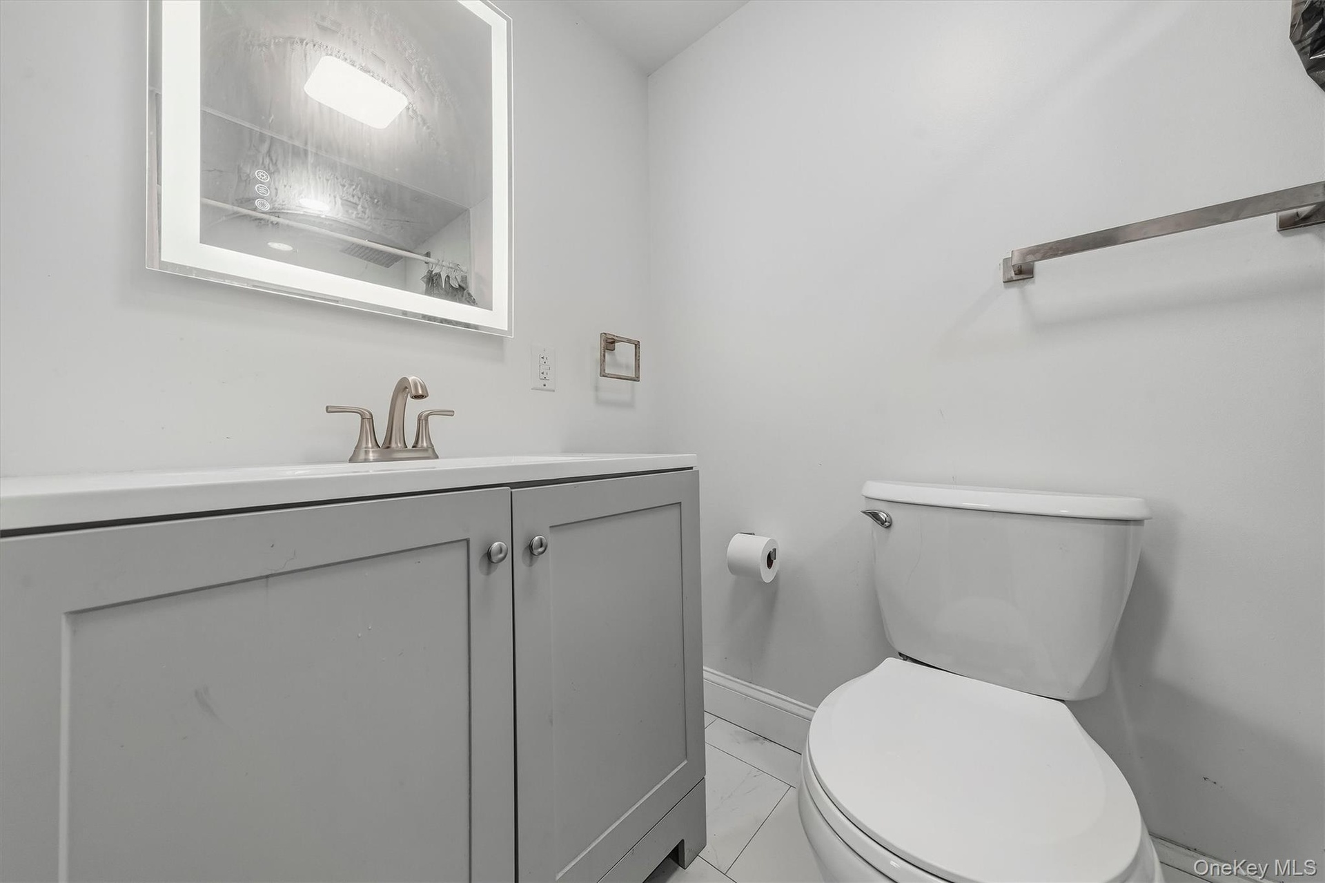 Lighted vanity mirror, gray vanity with cabinet storage, white countertop, modern brushed nickel faucet, and white tile flooring