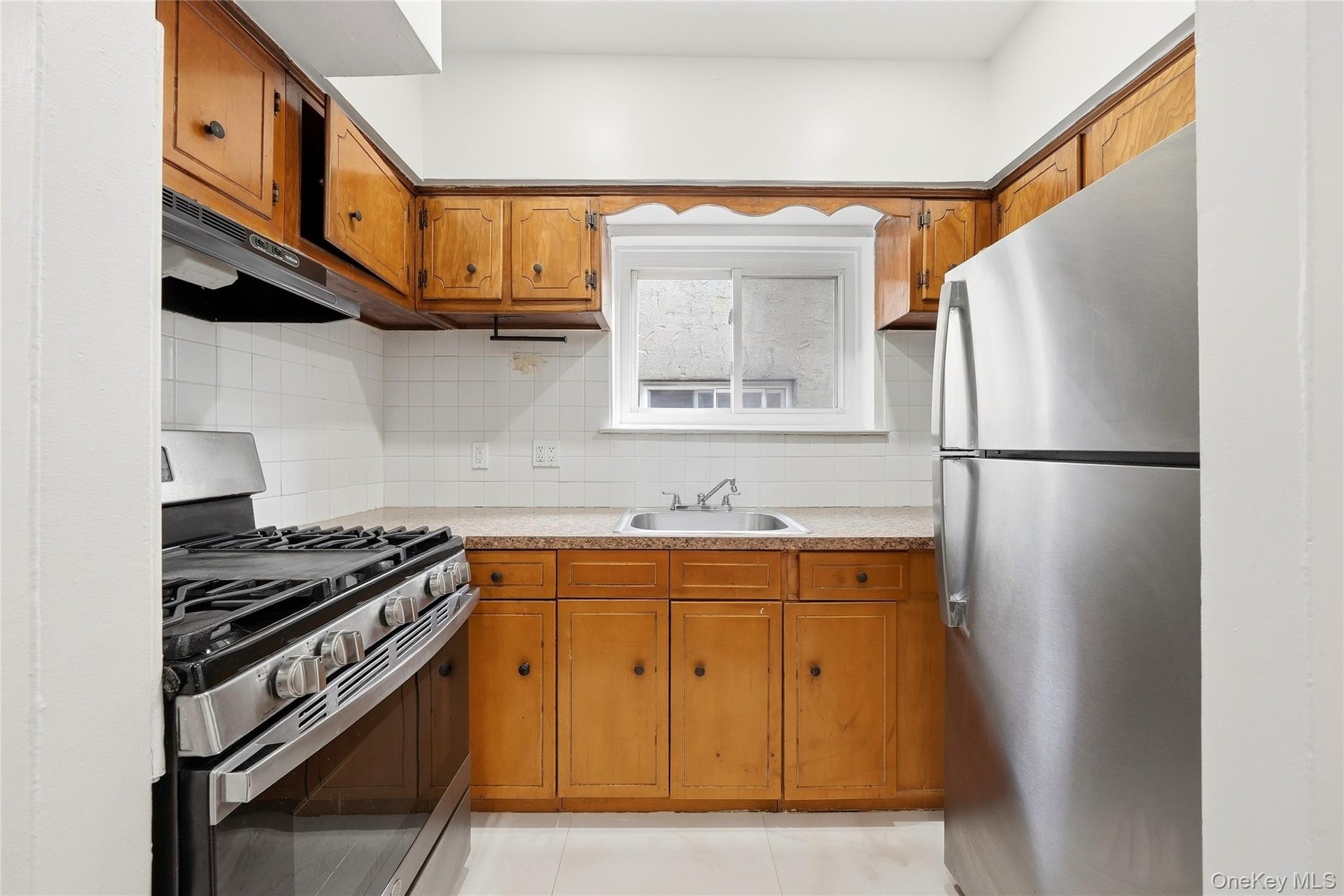 Kitchen featuring wood cabinetry, white tile backsplash, stainless steel range with overhead ventilation, and a stainless steel refrigerator