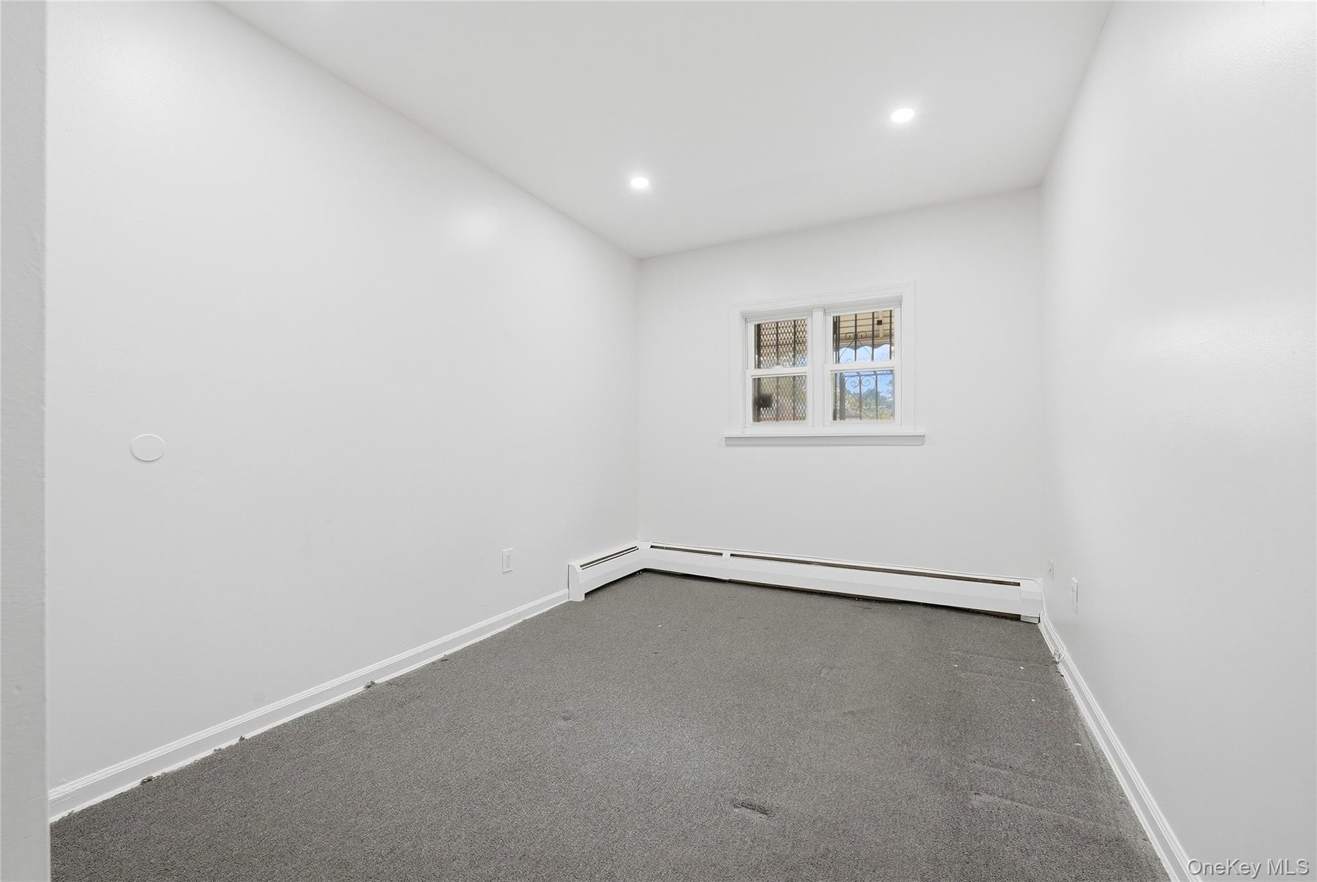 Recessed lighting illuminates a neutral painted interior with a window featuring security bars and baseboard heating