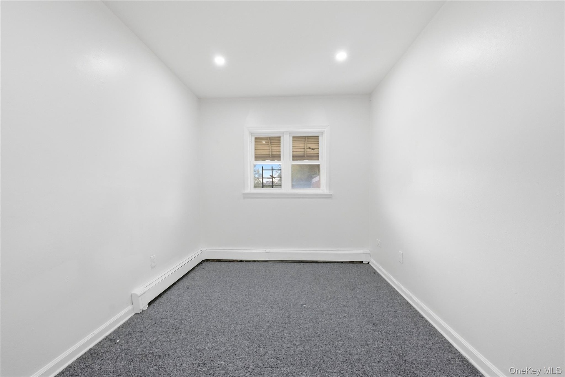 Recessed ceiling lighting, white painted walls, single window with blinds, baseboard heating unit, and gray carpeted flooring