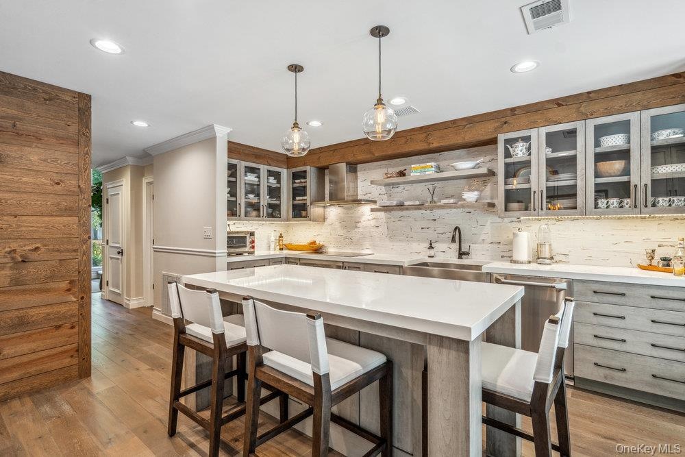 Kitchen featuring a large island with white countertops, wood-finish flooring, exposed wood beam accents, modern pendant lighting, and glass-front upp