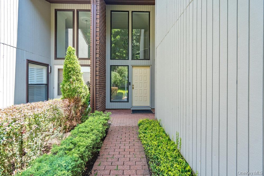 Brick paver pathway with manicured landscaping, vertical panel siding, and a paneled entry door