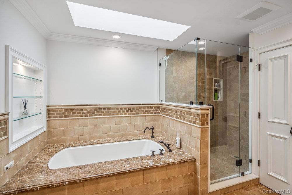 Soaking tub with a broad deck, enclosed shower with glass panels, recessed wall shelving with glass shelves, skylight, and crown molding