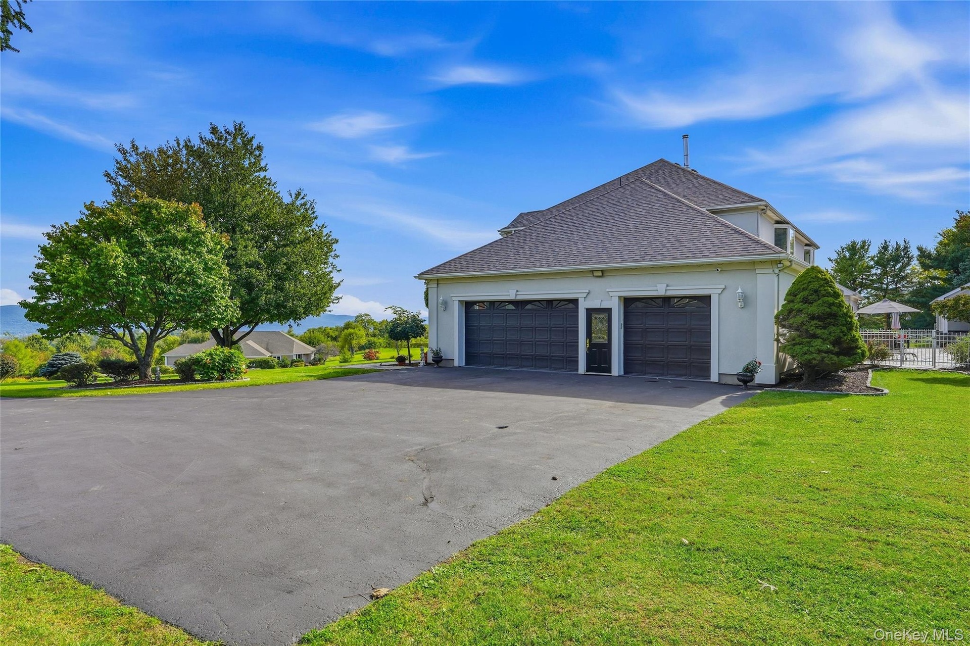 Three-car garage with black doors, paved driveway, expansive green lawn, mature trees, and a distant mountain backdrop