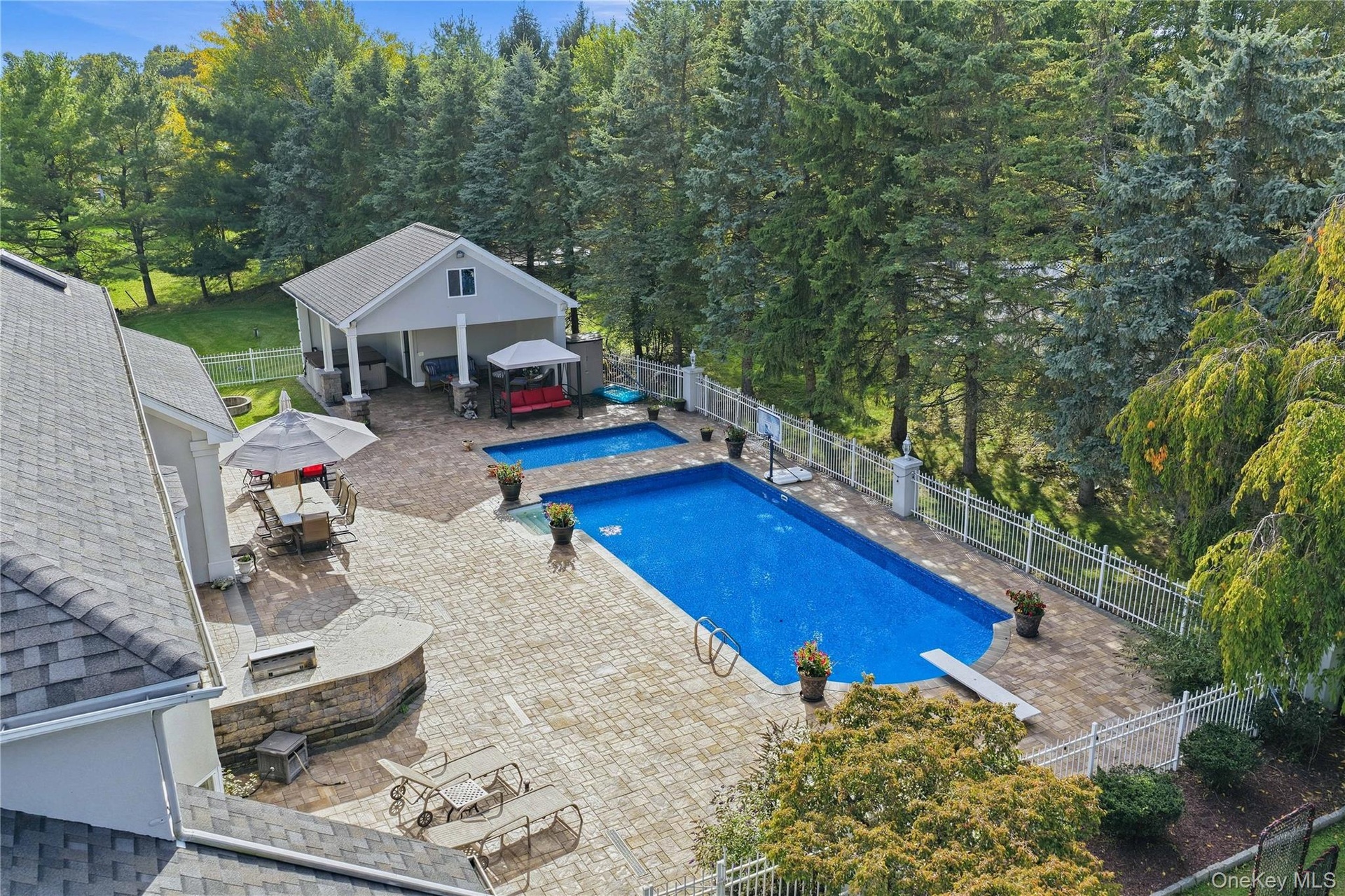 Expansive paver patio featuring a built-in outdoor kitchen, two swimming pools, and a pool house with covered outdoor space
