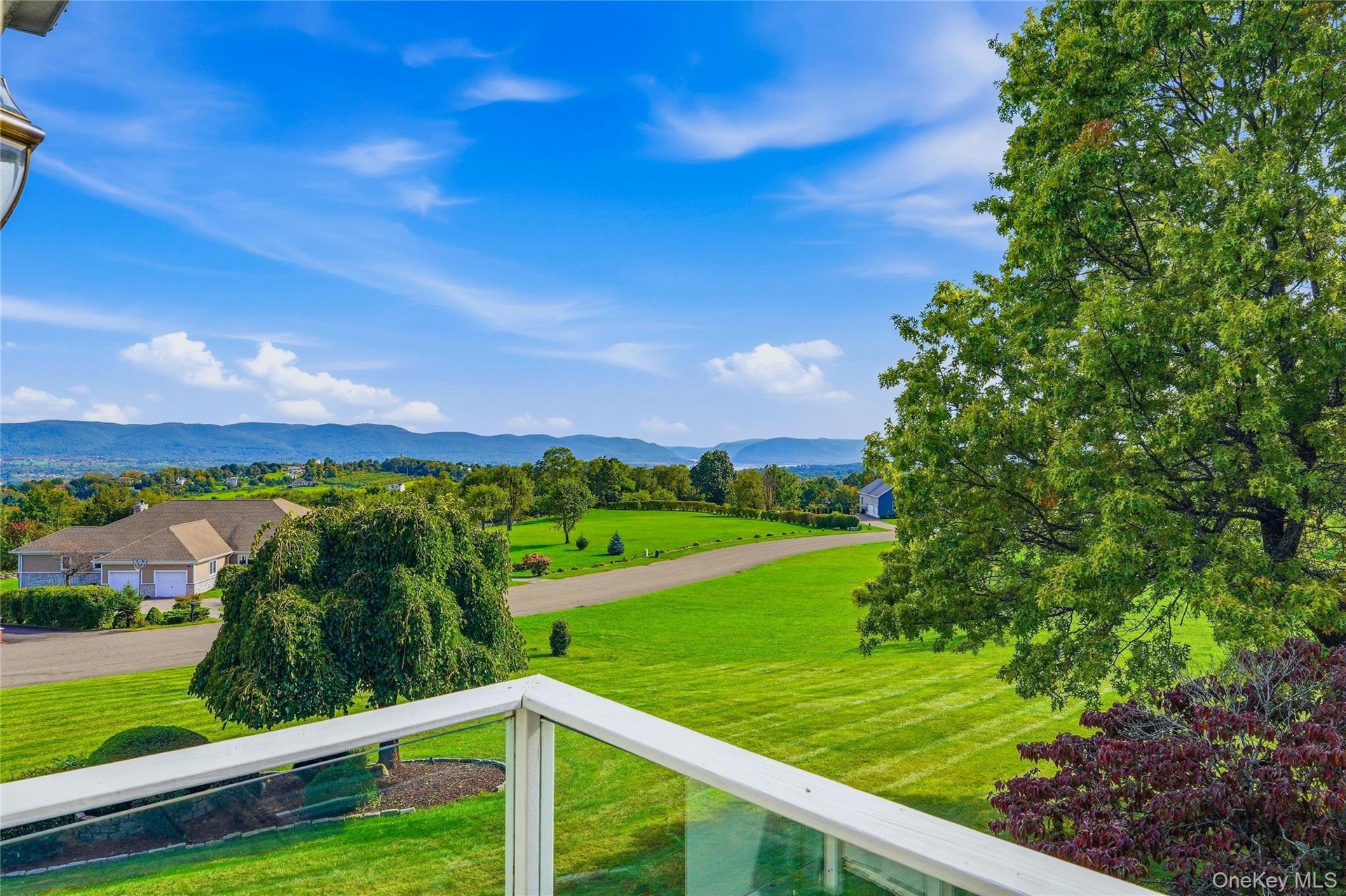 Balcony with glass railing, overlooking expansive landscape featuring rolling green hills, distant mountains, established trees, Hudson River views