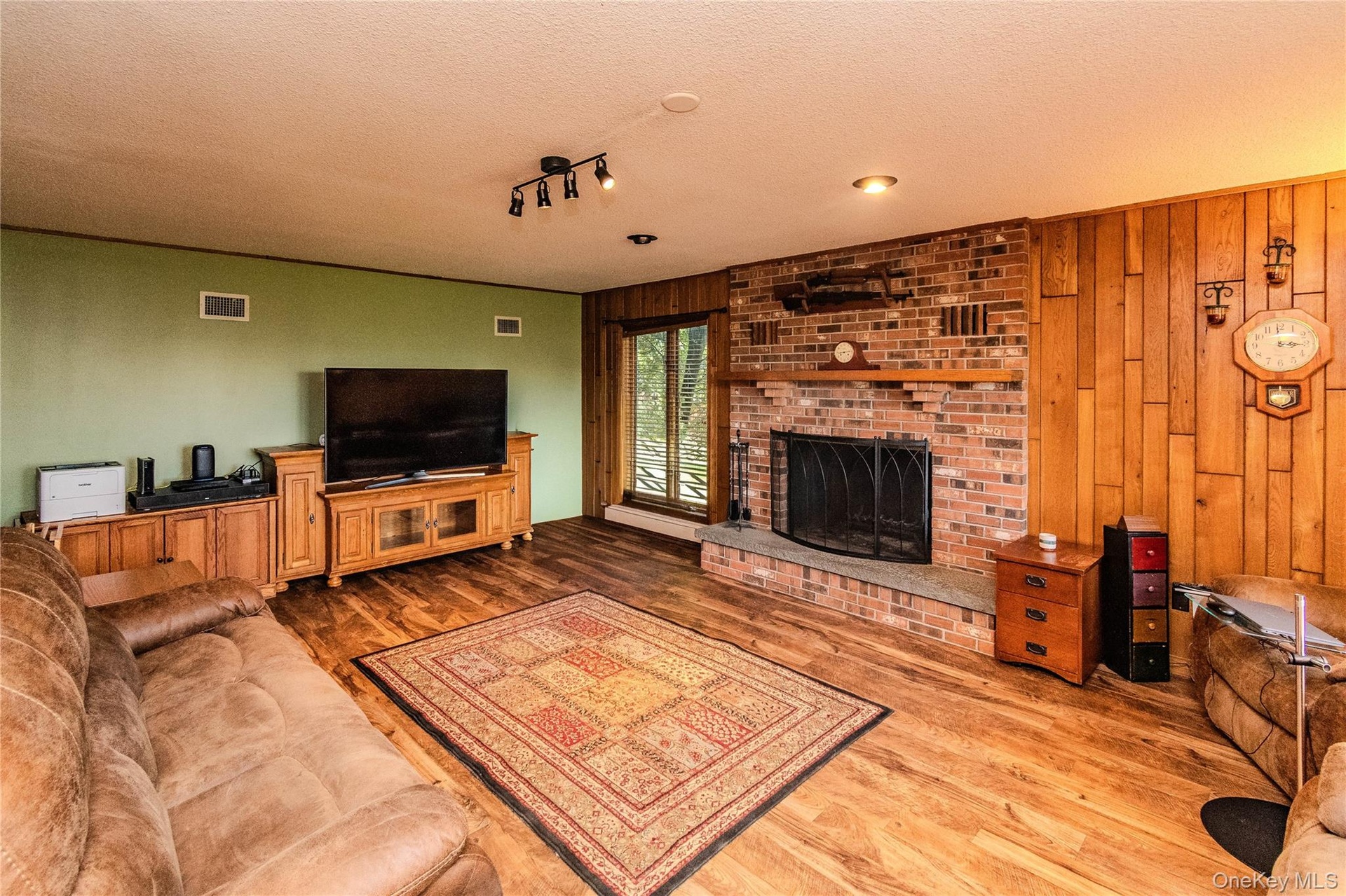 Wood-finish flooring throughout, featuring a brick fireplace with a stone hearth, wood paneling, and a large window