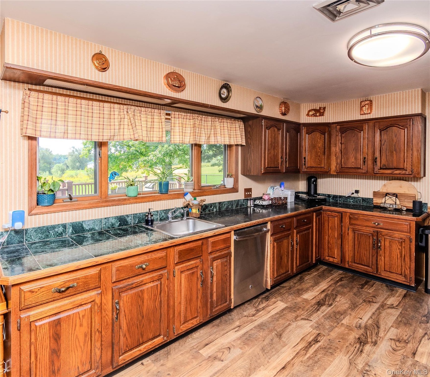 Wood-finish flooring, wood cabinetry, tile countertops, stainless steel sink, and stainless steel dishwasher