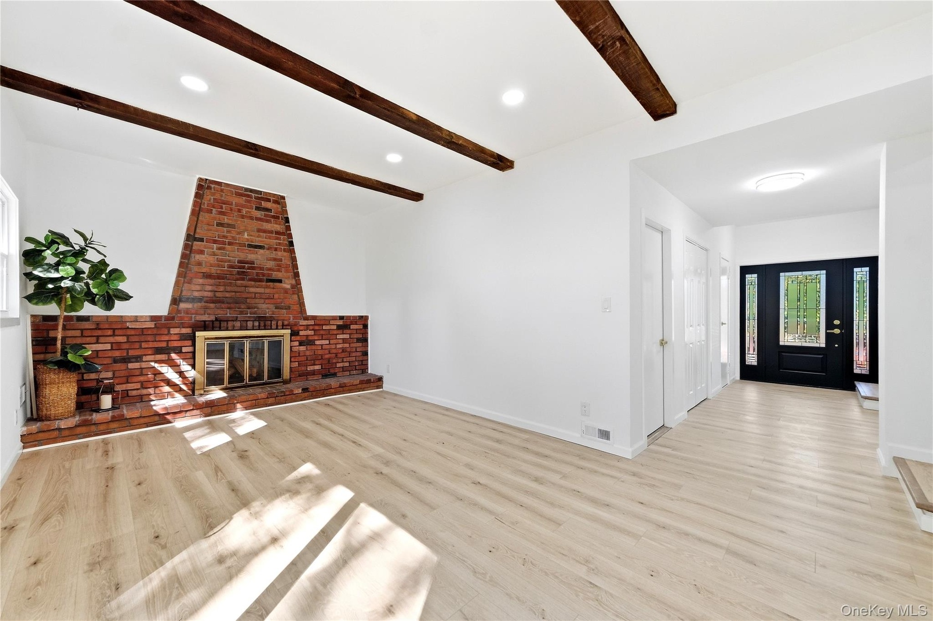 Exposed wood beams, recessed lighting, brick fireplace with metal insert, wood-finish flooring, and a dark-paneled entry door with decorative glass in