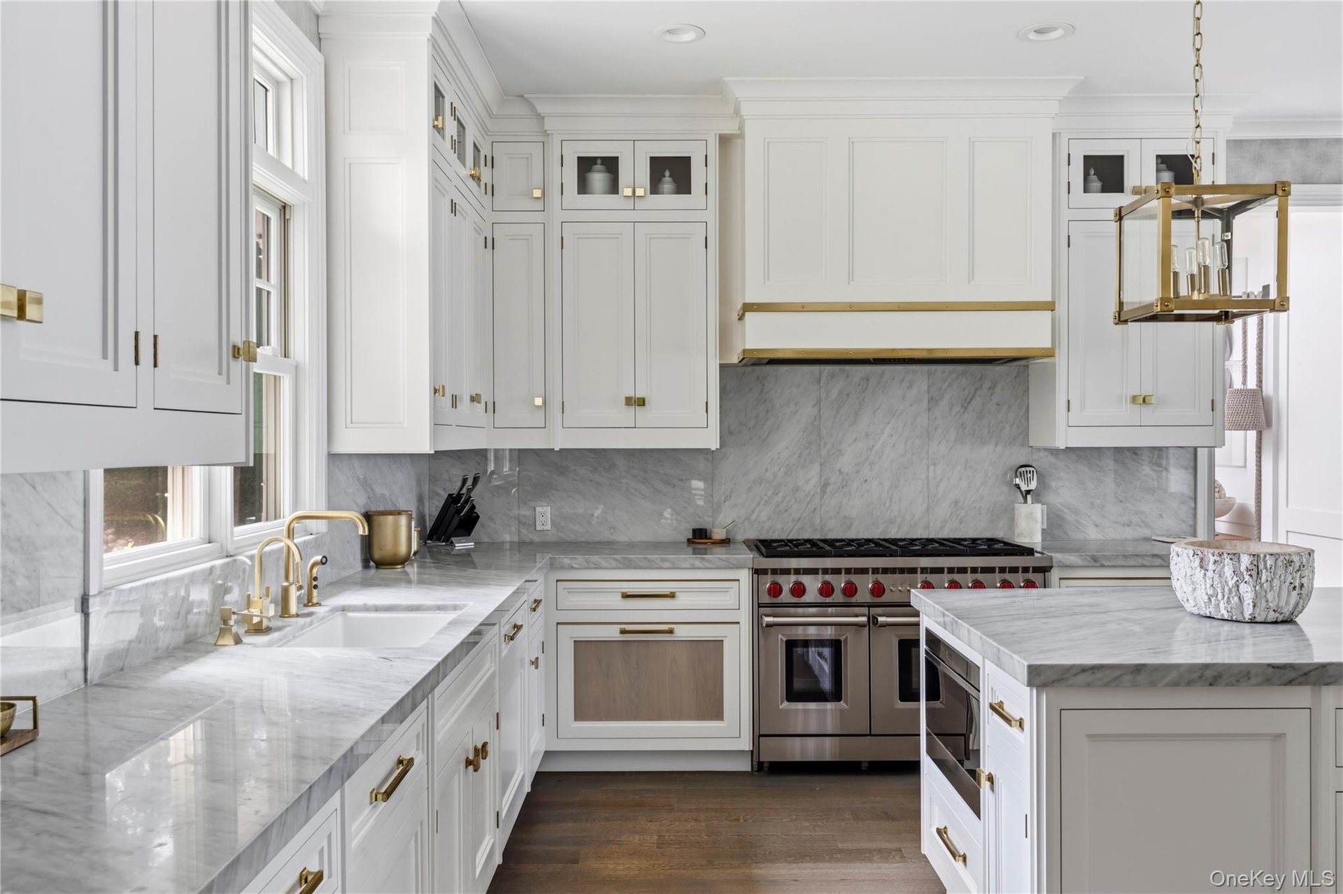 White cabinetry with gold-tone hardware, marble countertops and backsplash, stainless steel range, built-in microwave, and wood floors.