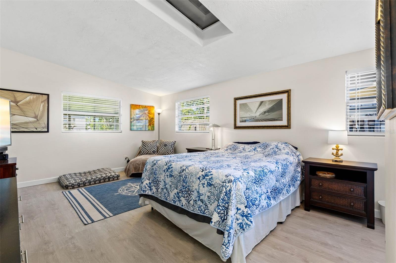 The generously sized primary bedroom feels bright and airy with multiple windows, a skylight overhead, neutral tones, and luxury vinyl plank flooring 