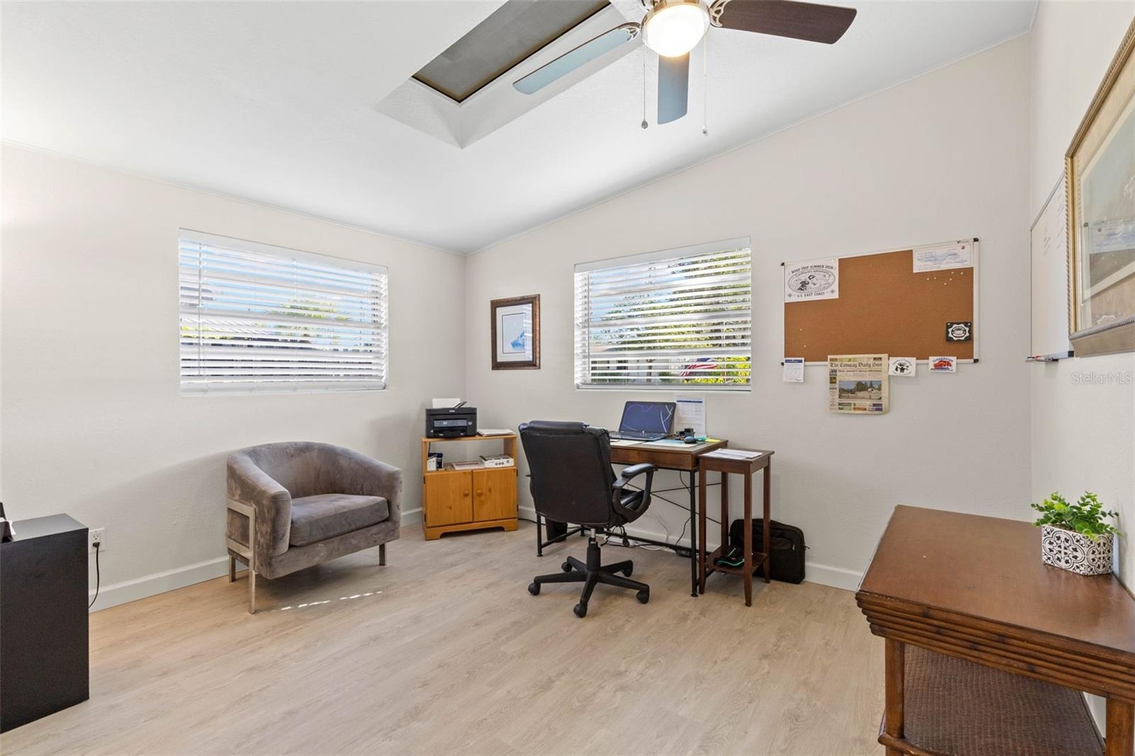 A bright secondary bedroom, currently used as a home office, offers two windows, a skylight, ceiling fan, neutral tones, and luxury vinyl plank floori