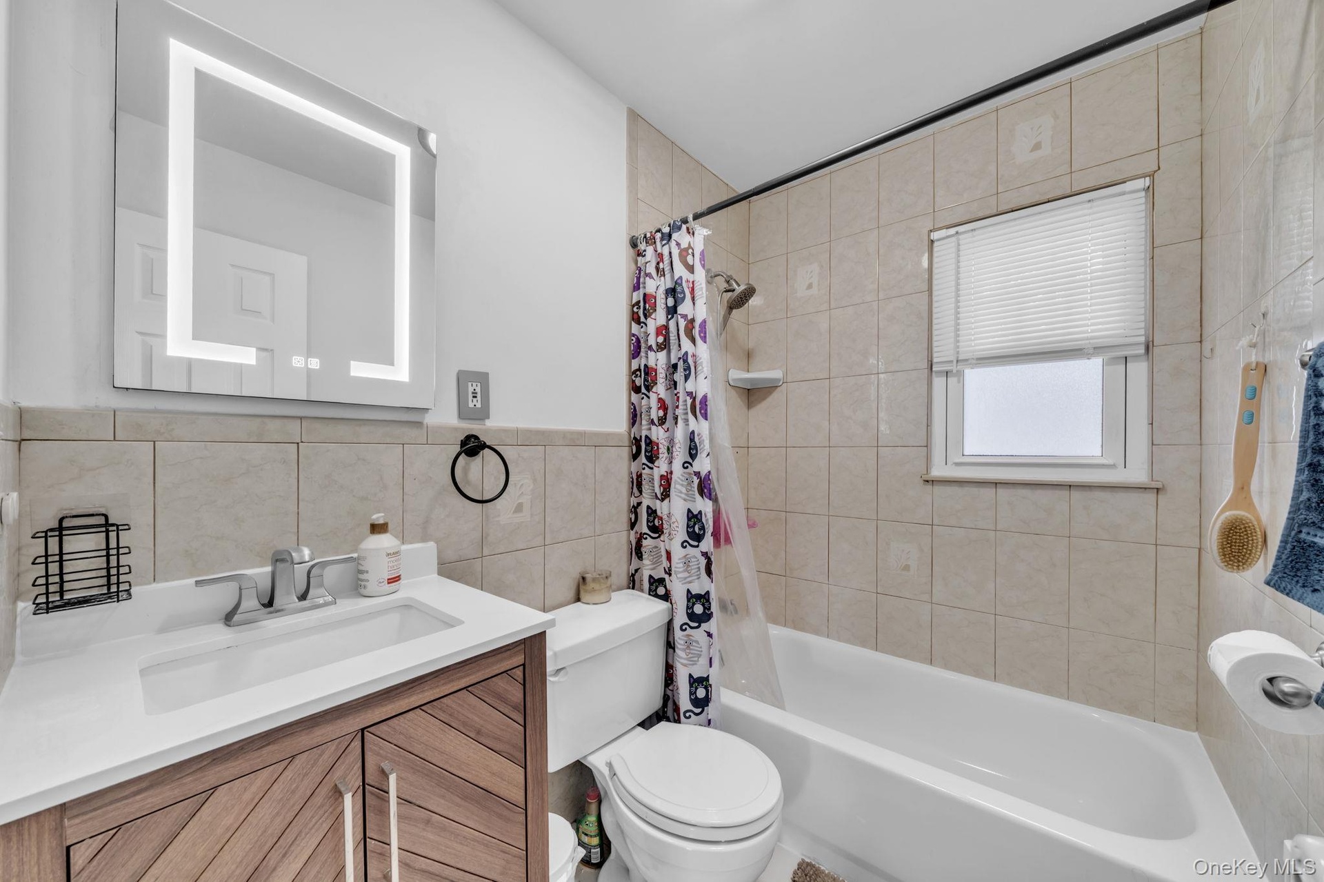 Illuminated vanity mirror, wood-finish vanity with an integrated sink, full bathtub with a shower fixture, and tiled walls