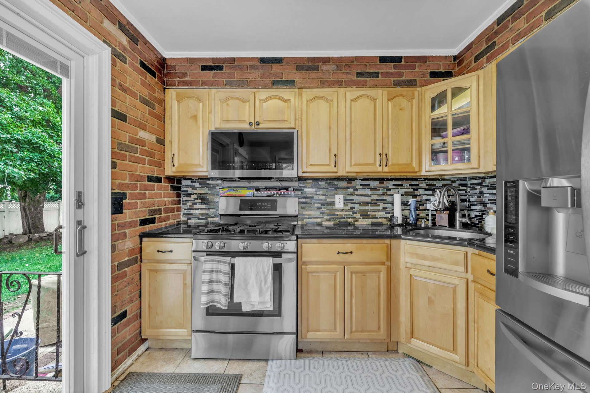 Kitchen featuring light wood cabinetry, a mosaic tile backsplash, black countertops, stainless steel appliances, and brick-patterned wall accents