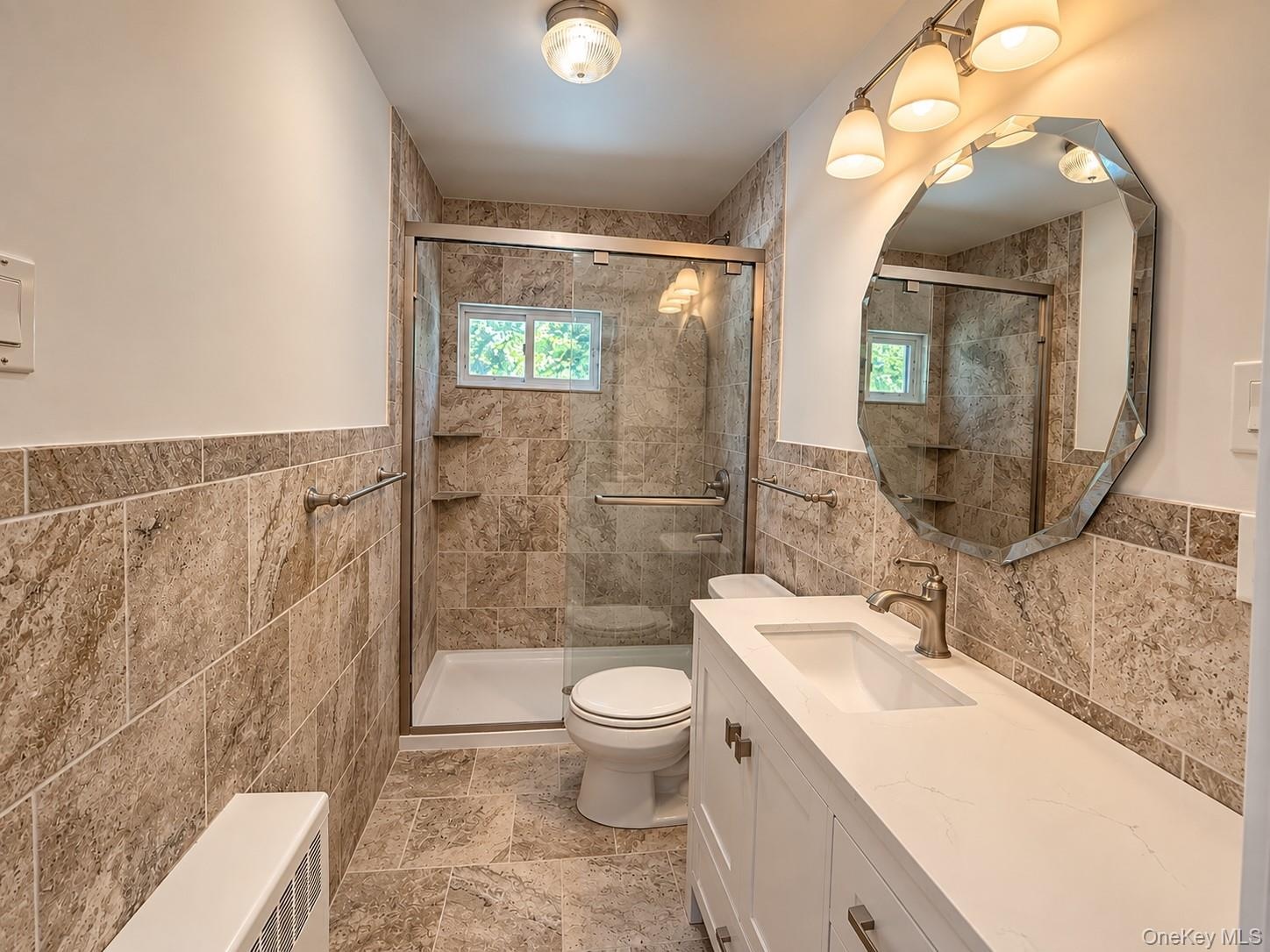 Tiled flooring and wainscoting, glass-enclosed shower with window, white vanity with integrated sink, contemporary vanity lighting, and decorative mir