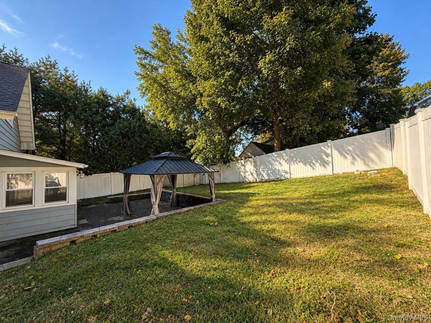 Expansive backyard featuring a gazebo with a dark-finish roof, a stone-block retaining wall, a paved patio area, and a white privacy fence