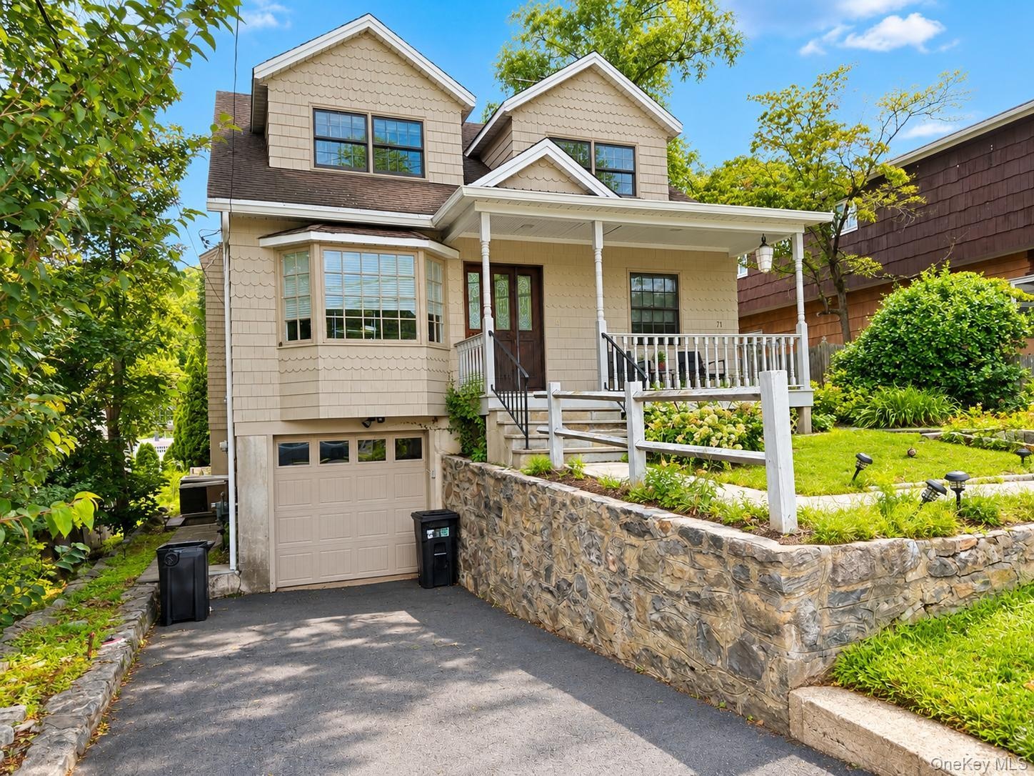 Gable roofline with dormer windows, bay window, front porch with white railings, integrated garage, and stone retaining wall