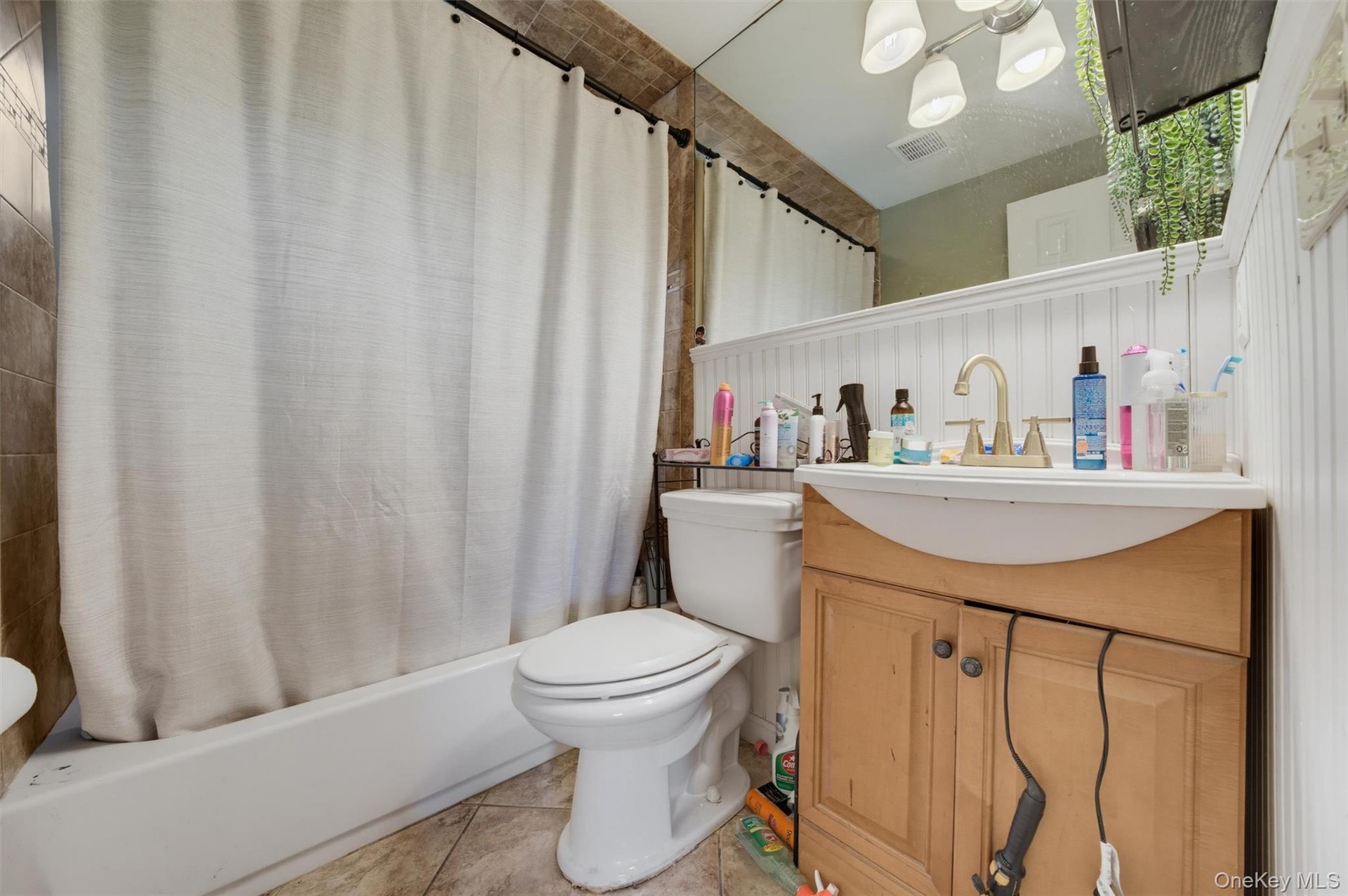 Beige tile wall and floor, white beadboard paneling, light wood-finish vanity, and a white bathtub