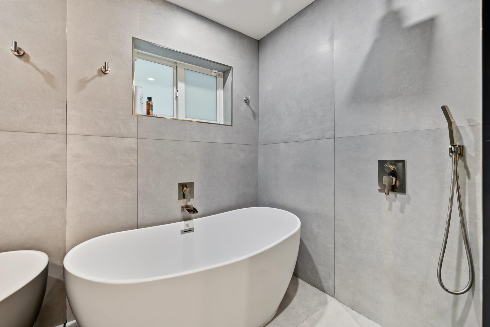 Freestanding soaking tub with brushed nickel fixtures, surrounded by large format wall tiling and a built-in window