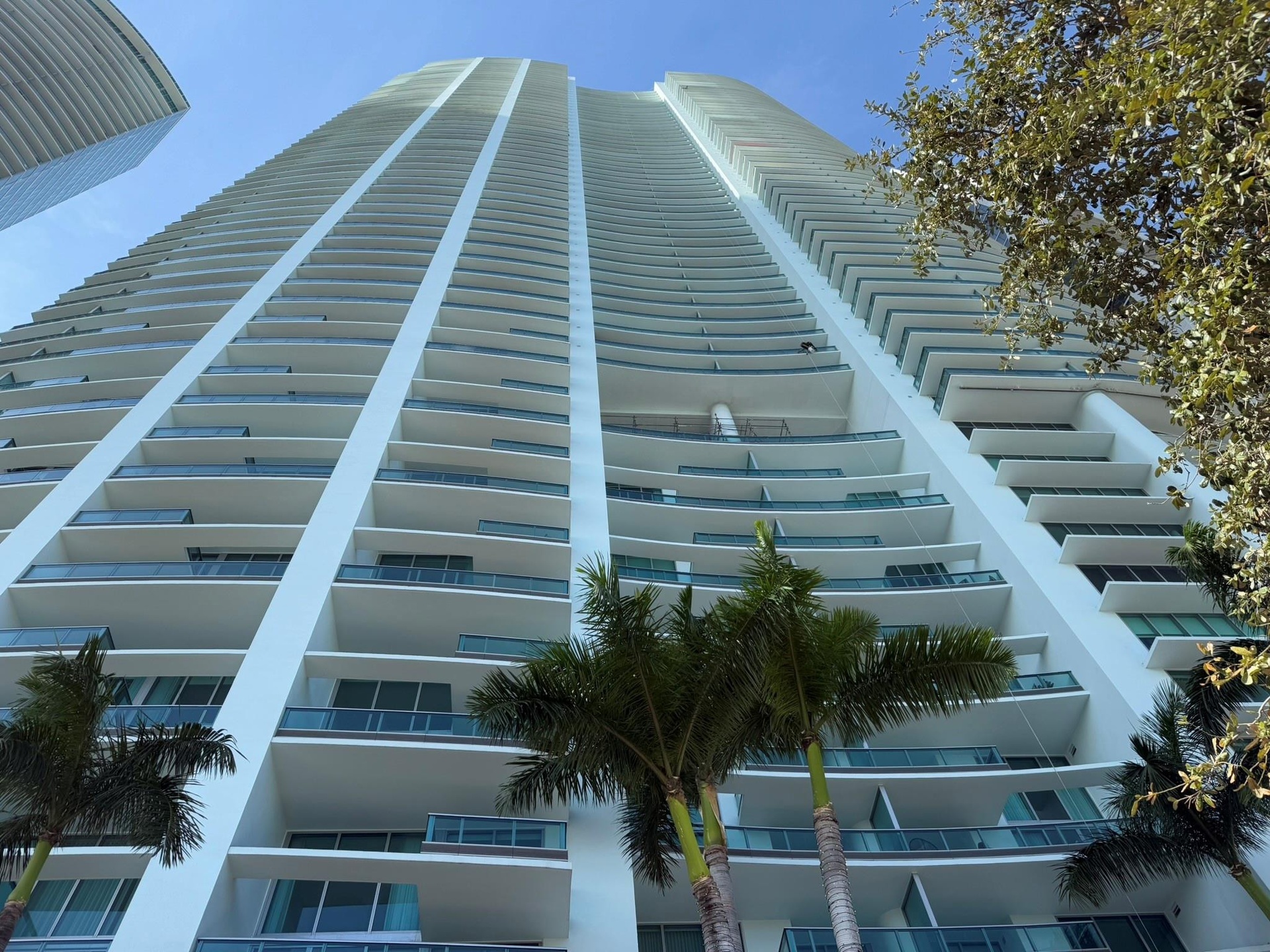 Contemporary high-rise building with a white exterior, multiple curved balconies, glass balcony railings, floor-to-ceiling windows, and surrounding pa