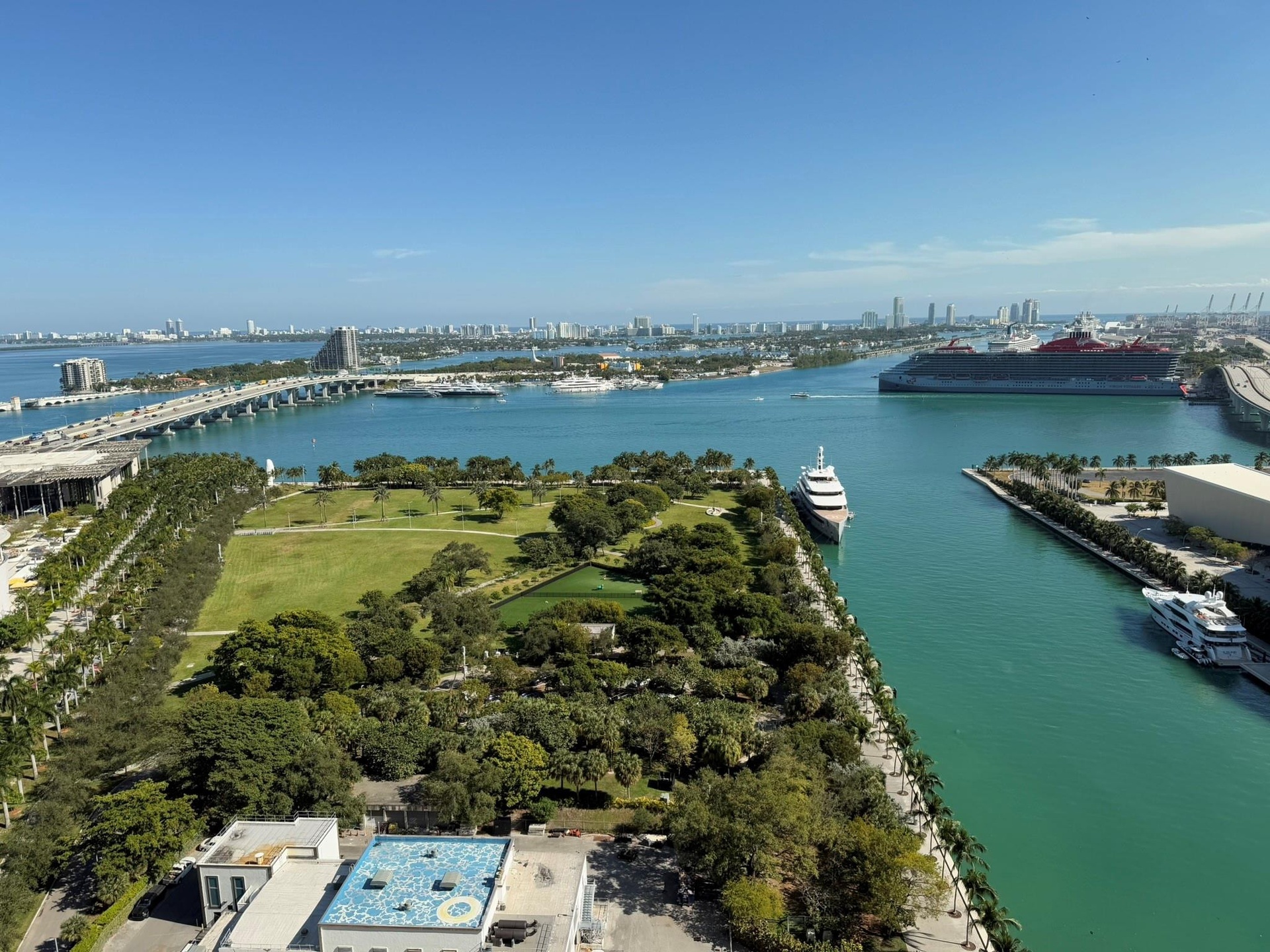 Expansive harbor and city skyline panorama featuring water views, a marine channel, lush parkland, and a distant bridge structure