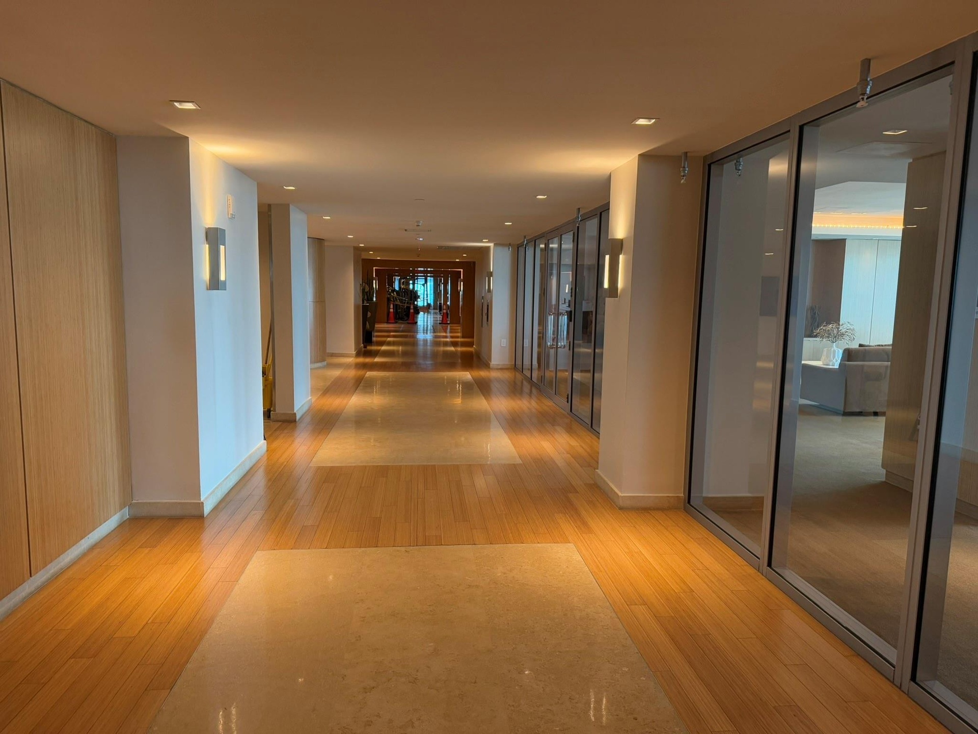 Expansive hallway featuring wood-finish flooring with alternating stone inlay, recessed ceiling lighting, and wall-mounted sconces