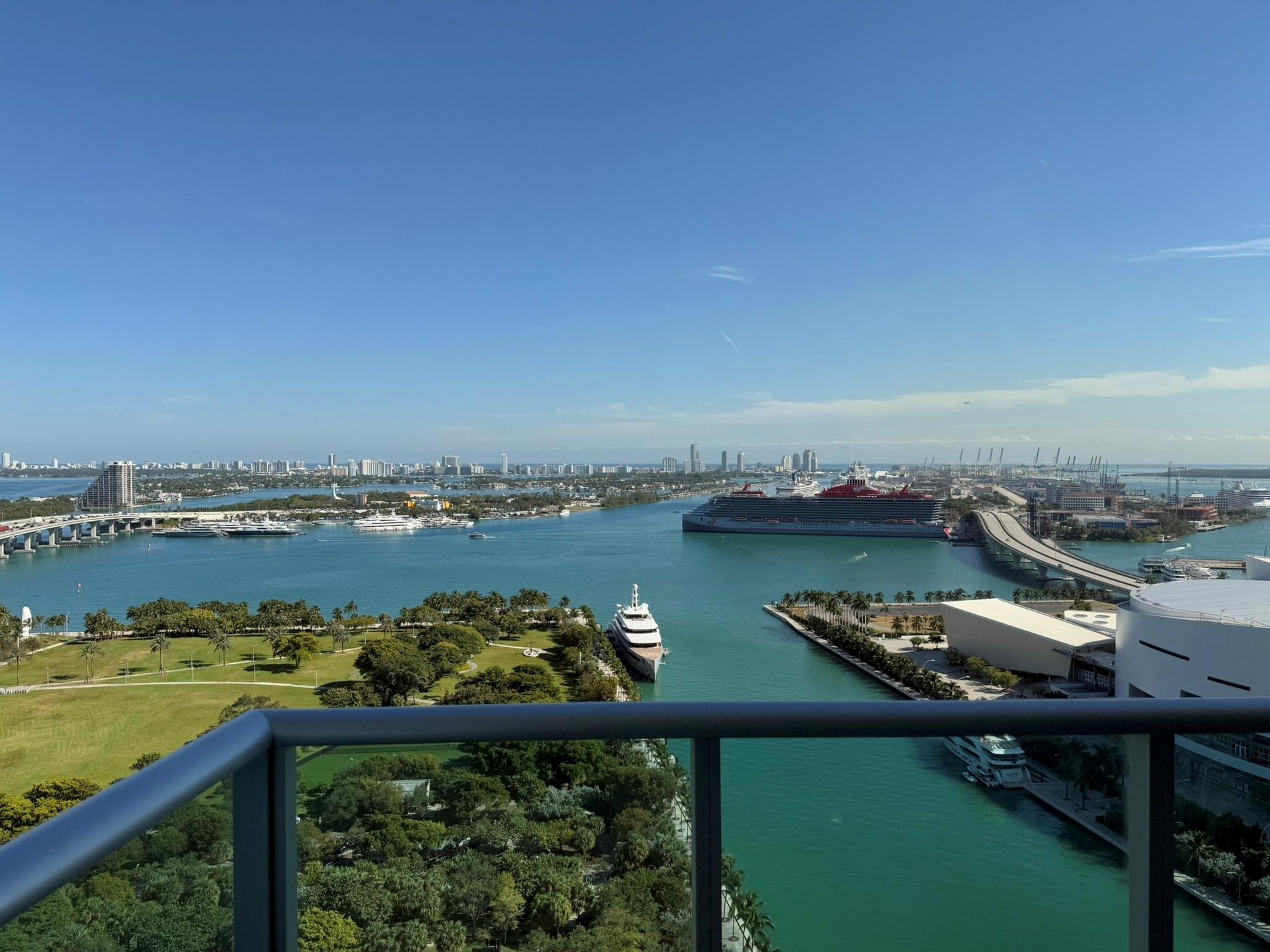 Expansive waterfront scenery featuring a cityscape skyline, open water, lush green parkland, a bridge, and a distant cruise ship