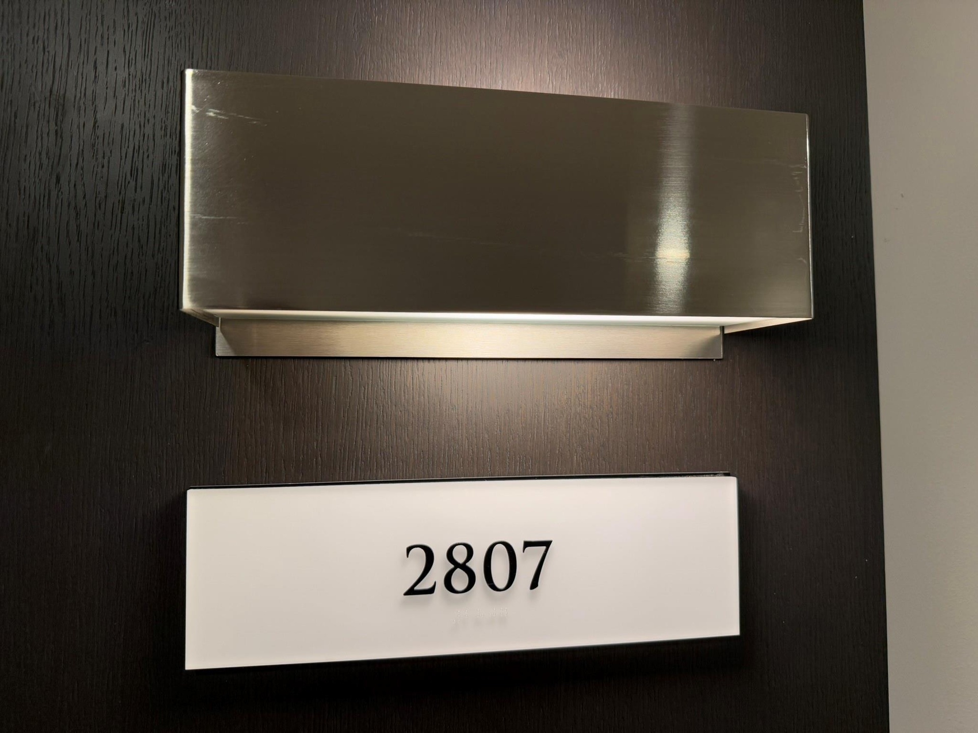 Dark wood-grain door with an illuminated brushed metal fixture above a numeric wall sign