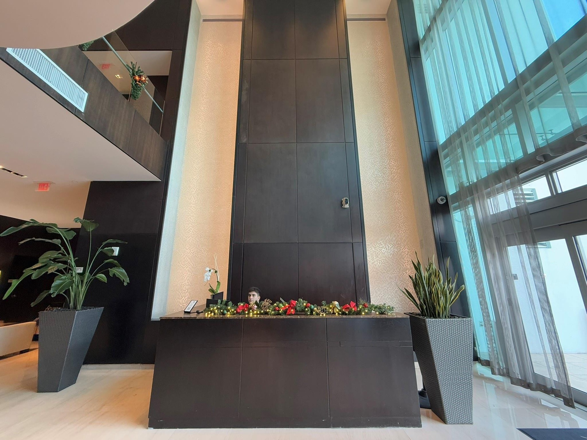 Two-story lobby featuring a dark wood-finish reception desk, light-toned tiled flooring, and a full-height glass window wall with sheer drapery