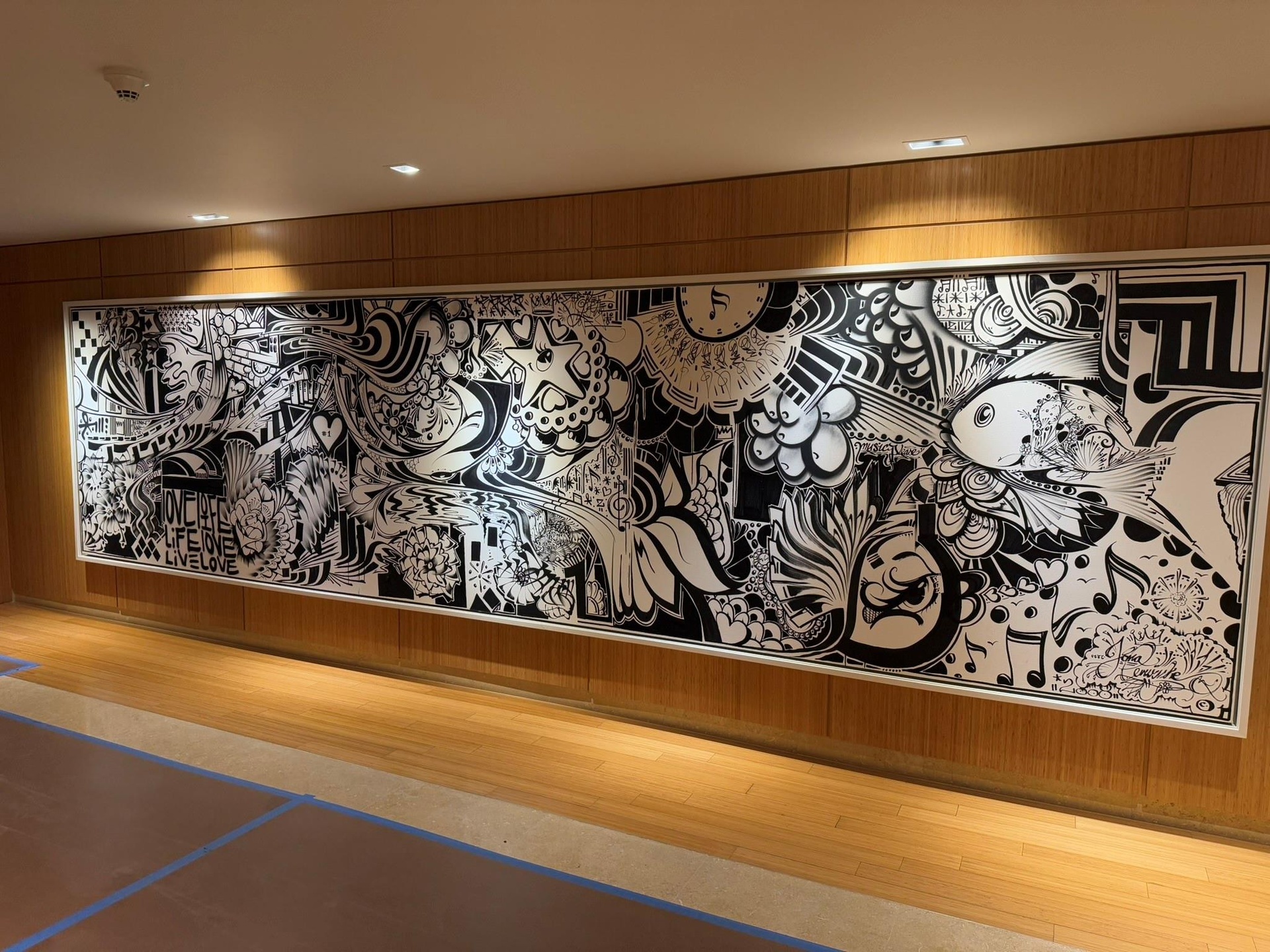 Large-scale black and white graphic artwork mounted on a wood-paneled wall, illuminated by integrated ceiling lighting